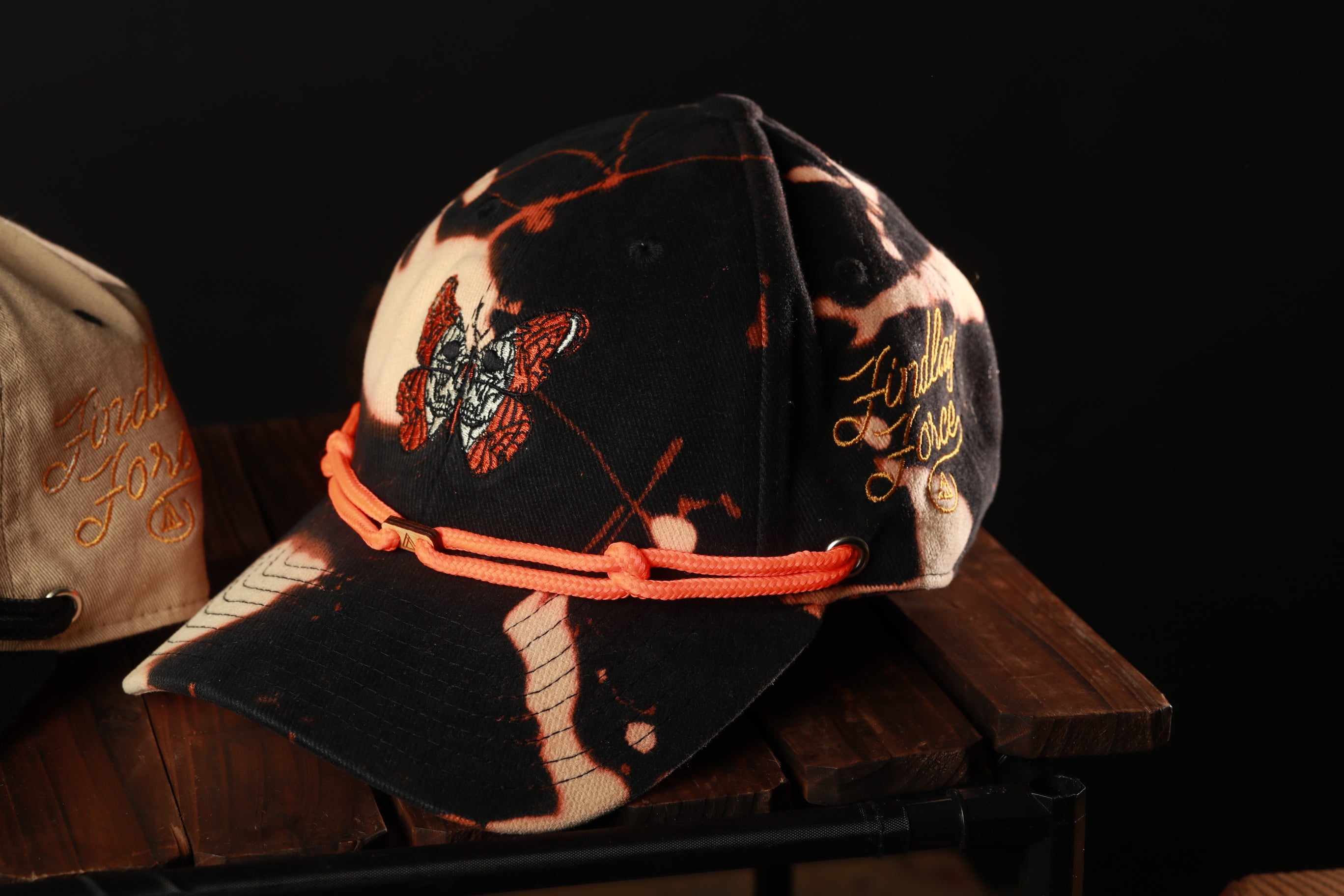 A black and orange baseball cap with a butterfly design, featuring a rope strap, is displayed on a wooden surface.