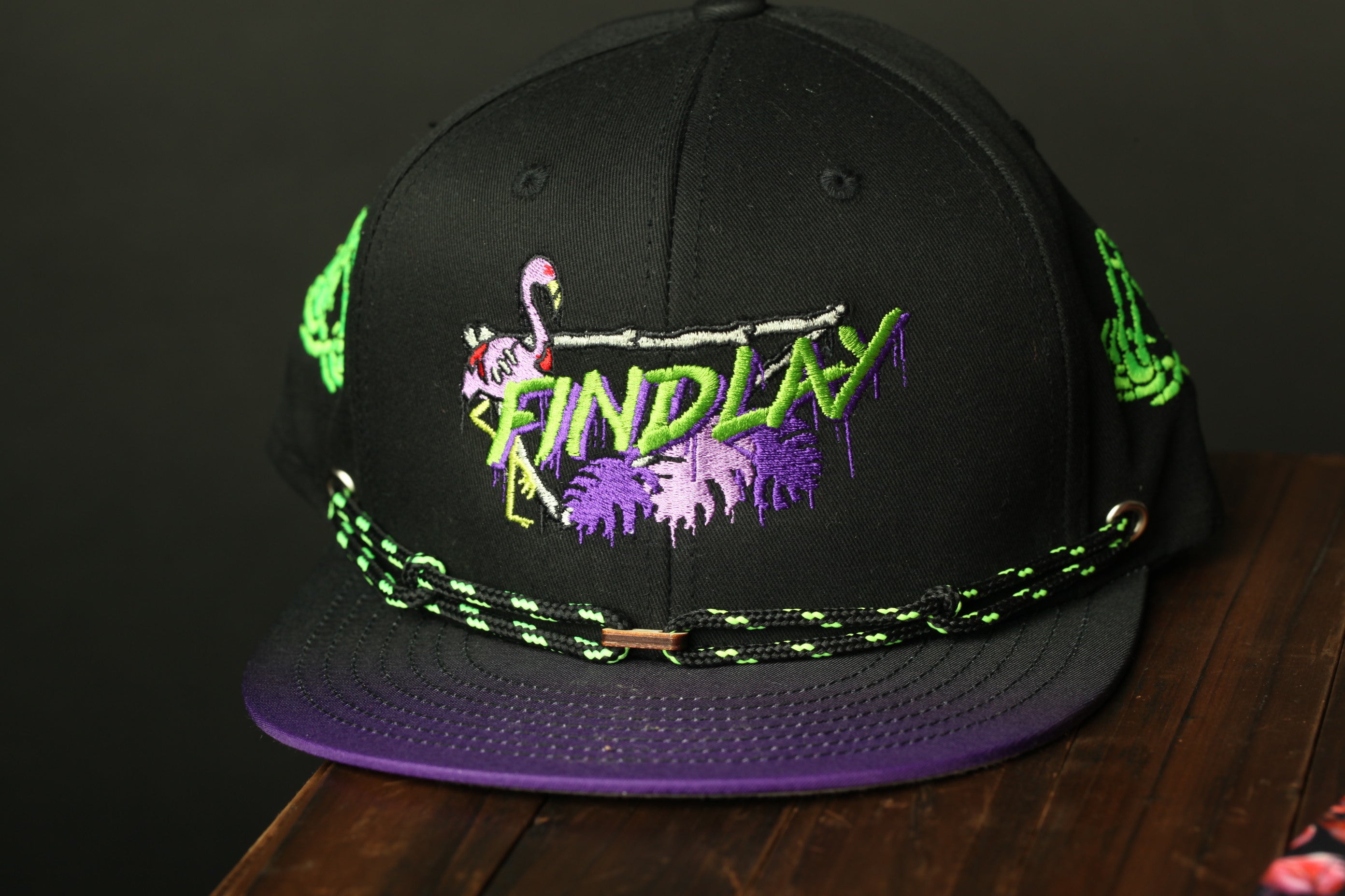 A black baseball cap with a purple brim and neon green and purple designs, including a flamingo and a skull.