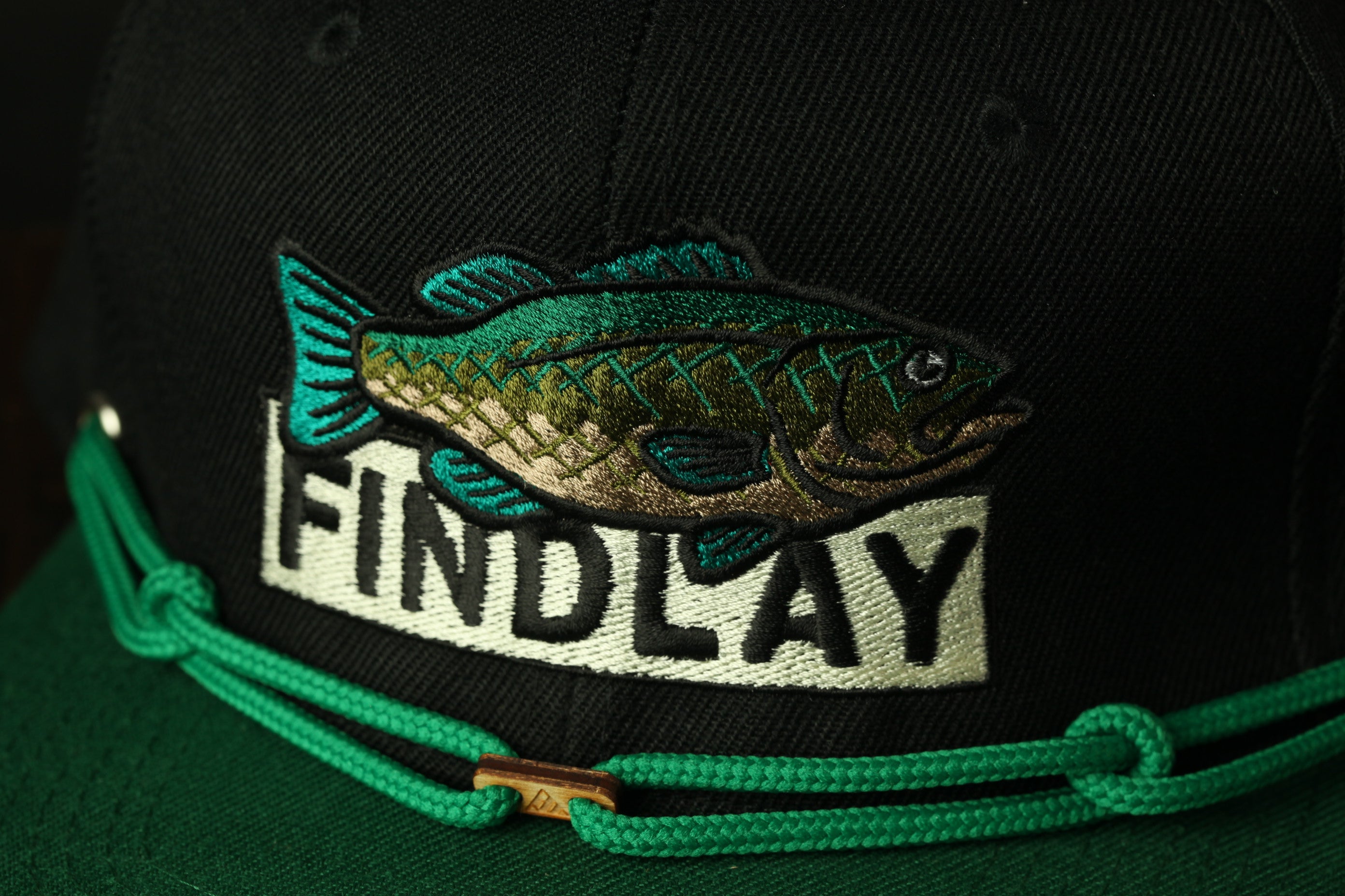 A close-up view of a black baseball cap with a fish embroidered on it, and the word "FINDLAY" printed on the front.