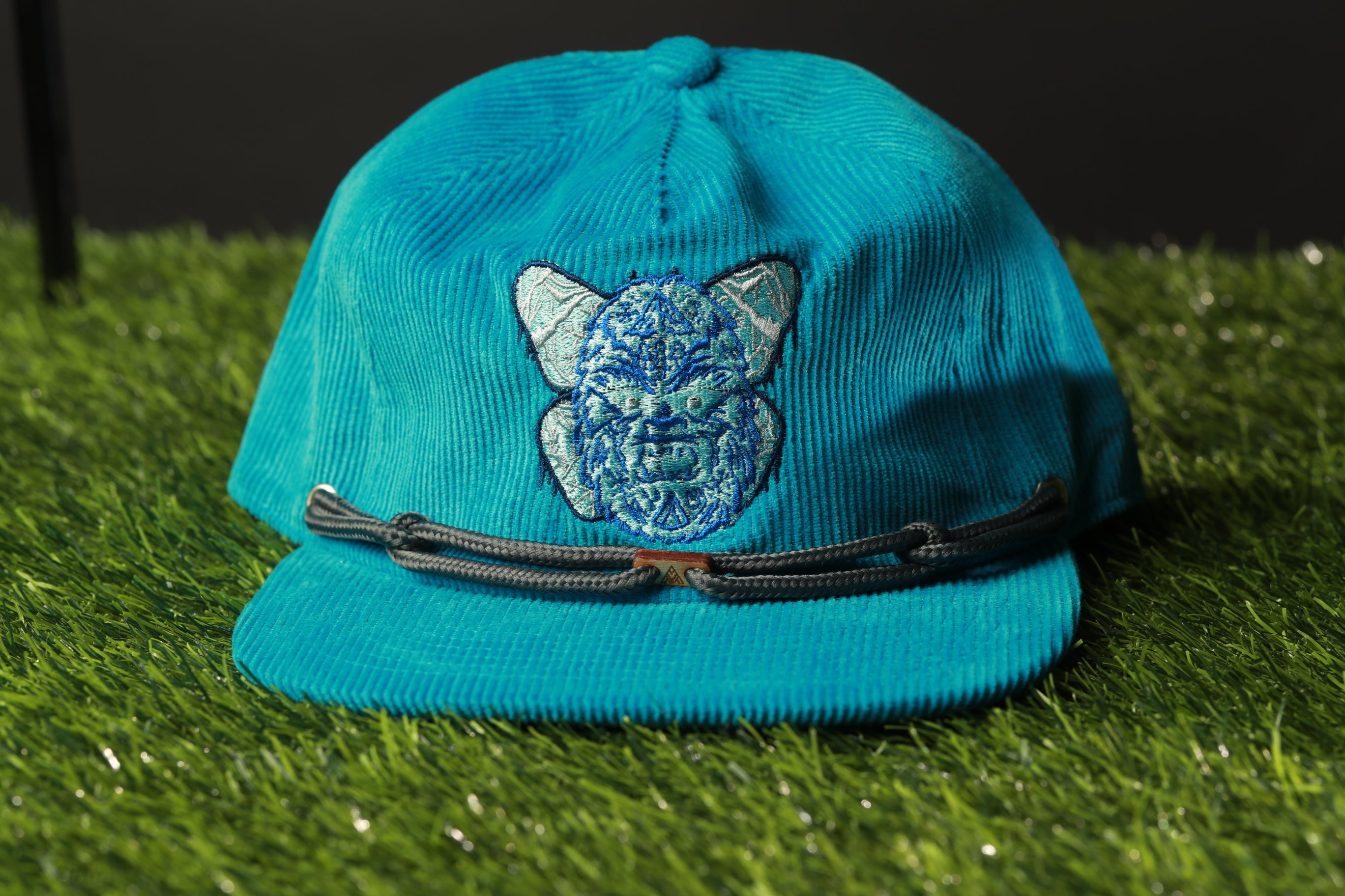 A turquoise baseball cap with a cat embroidered on it, placed on a grassy surface.