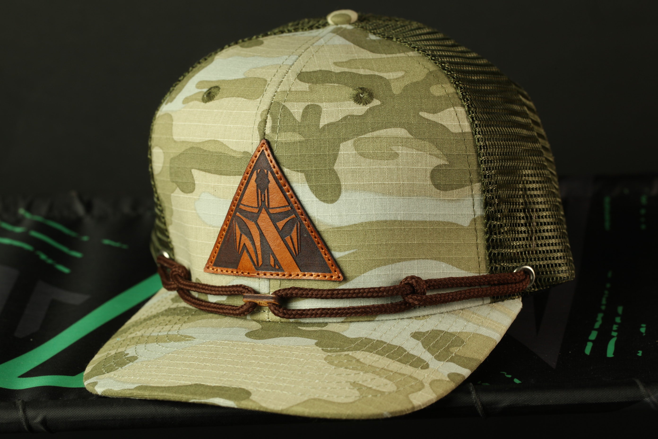 A camouflage baseball cap with a leather patch and a brown braided strap.