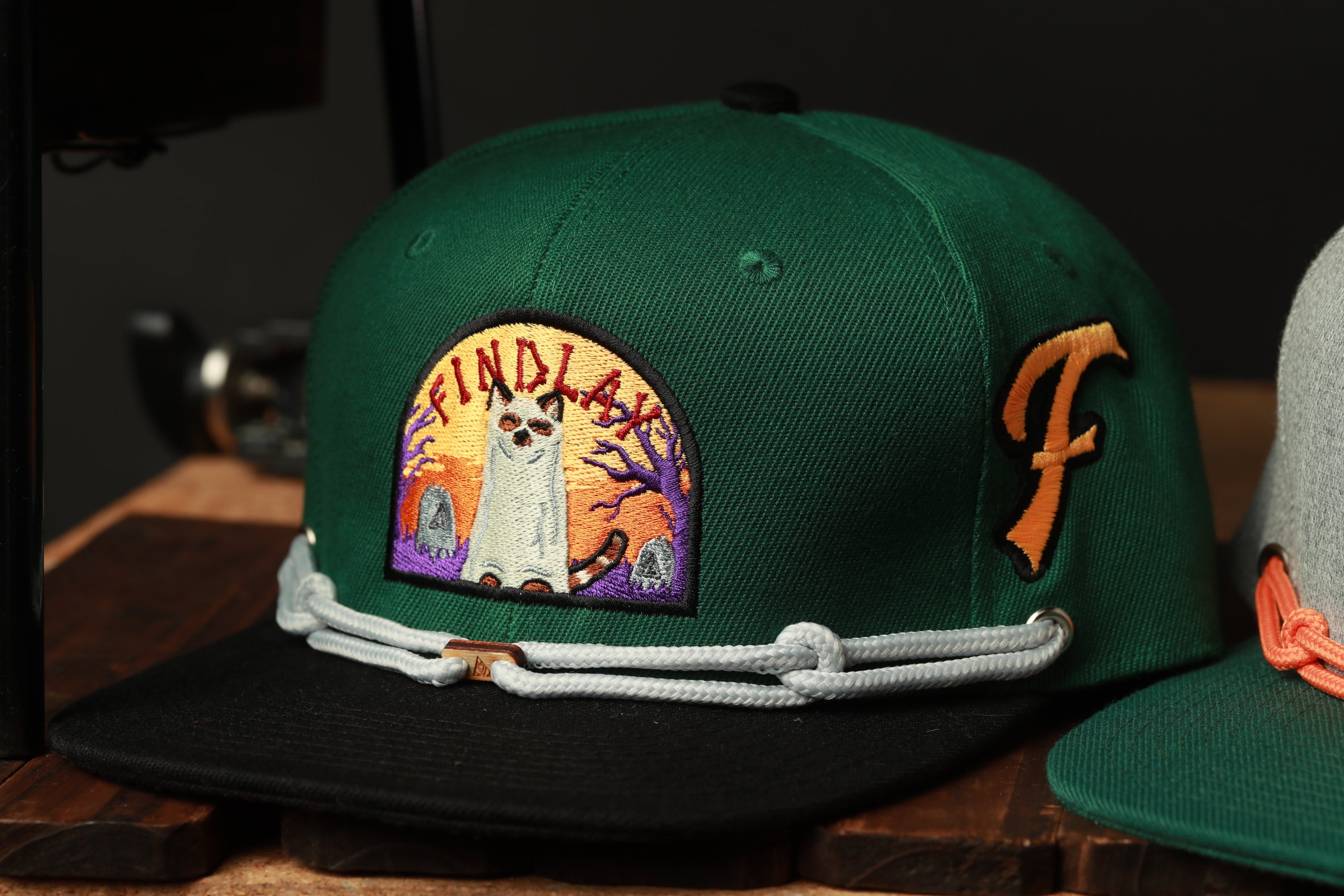 A green baseball cap with a cat embroidered on it, featuring a colorful design and a chain strap.