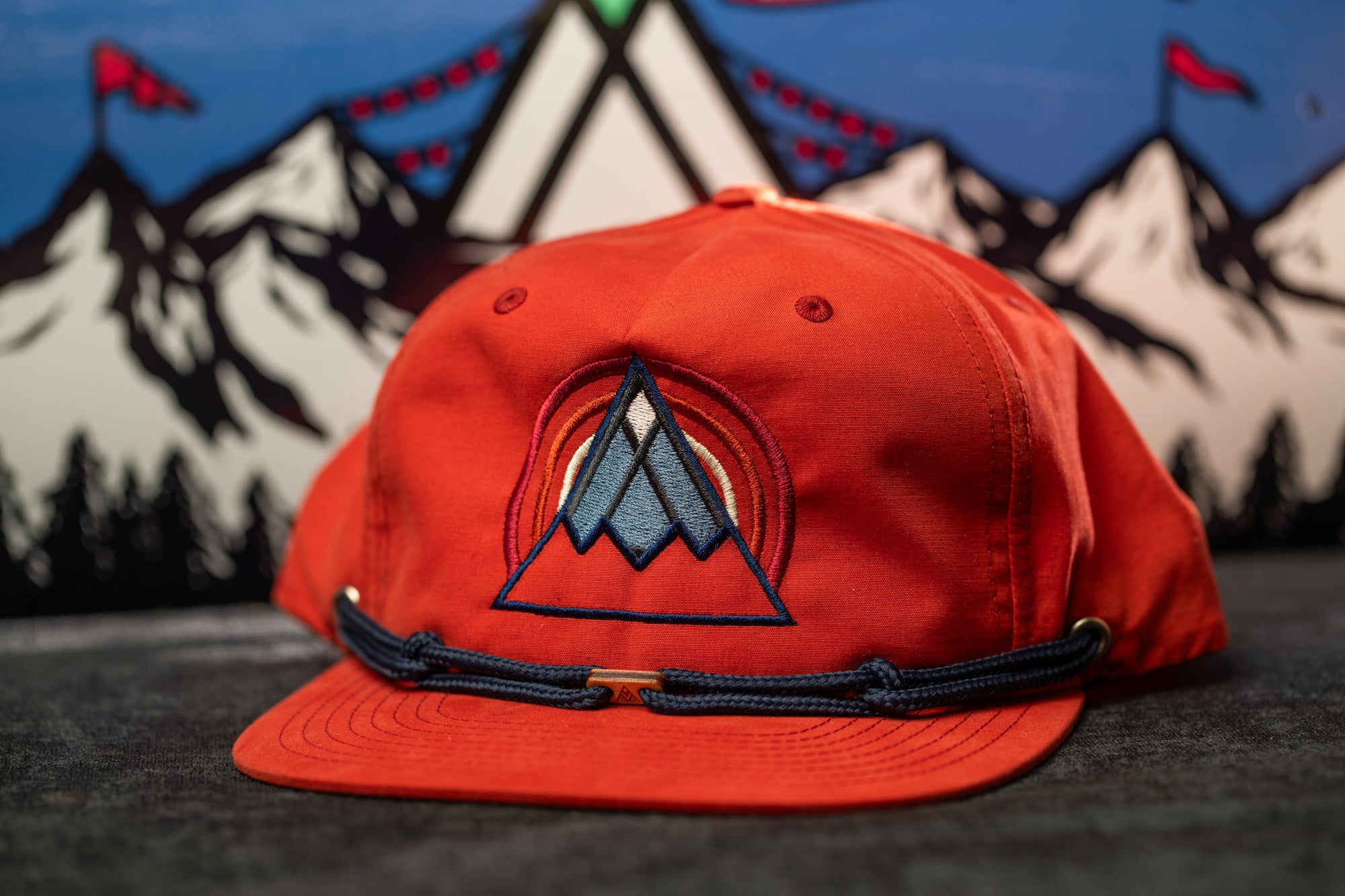 A red baseball cap with a mountain-themed design is displayed against a background of mountains and flags.