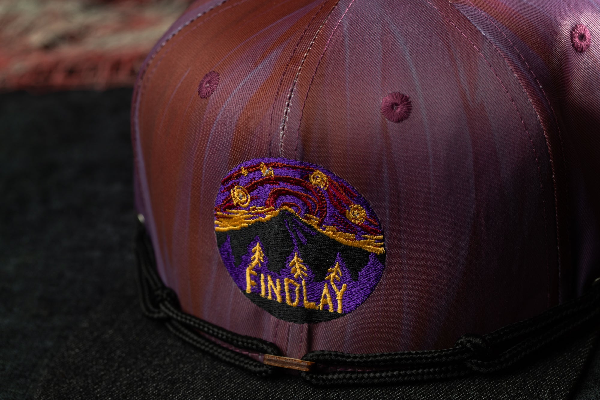 A close-up image of a purple baseball cap with a circular embroidered patch on the front, featuring a mountain landscape and the word "FINOLAY".