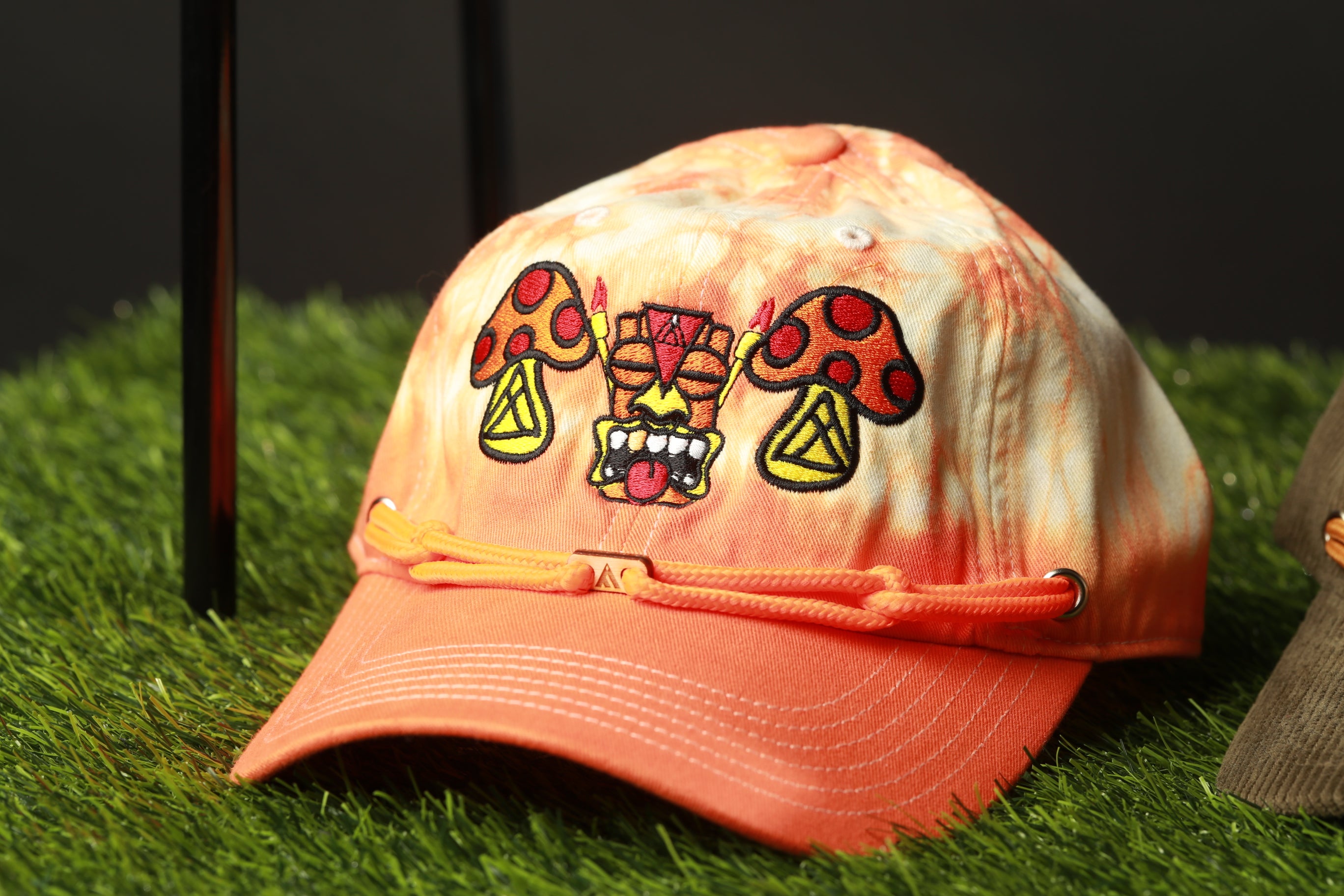 An orange baseball cap with a mushroom and tiki mask design on it, placed on a grassy surface.