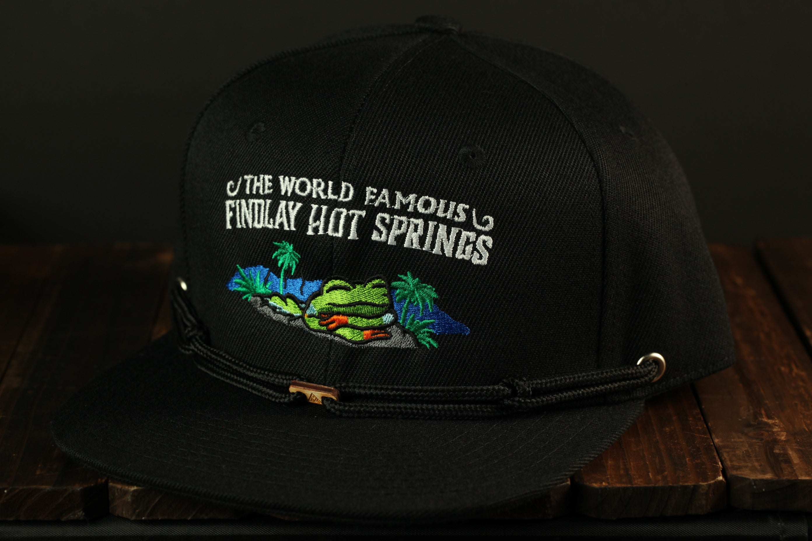 A black baseball cap with a printed design on the front, featuring text and an illustration of a tropical landscape.