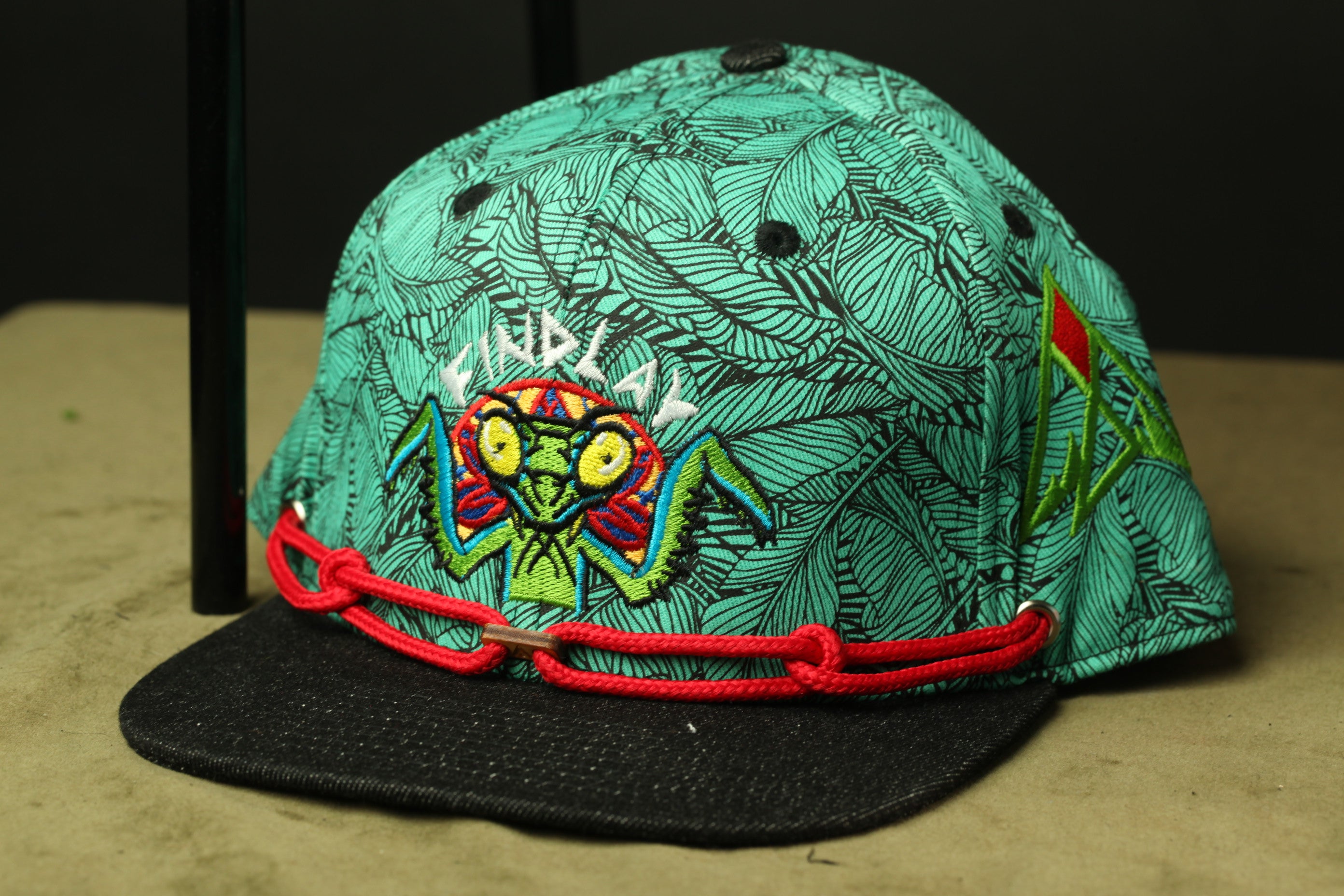 A colorful, patterned baseball cap with a black brim and red straps, featuring a unique design with a stylized insect or creature on the front.