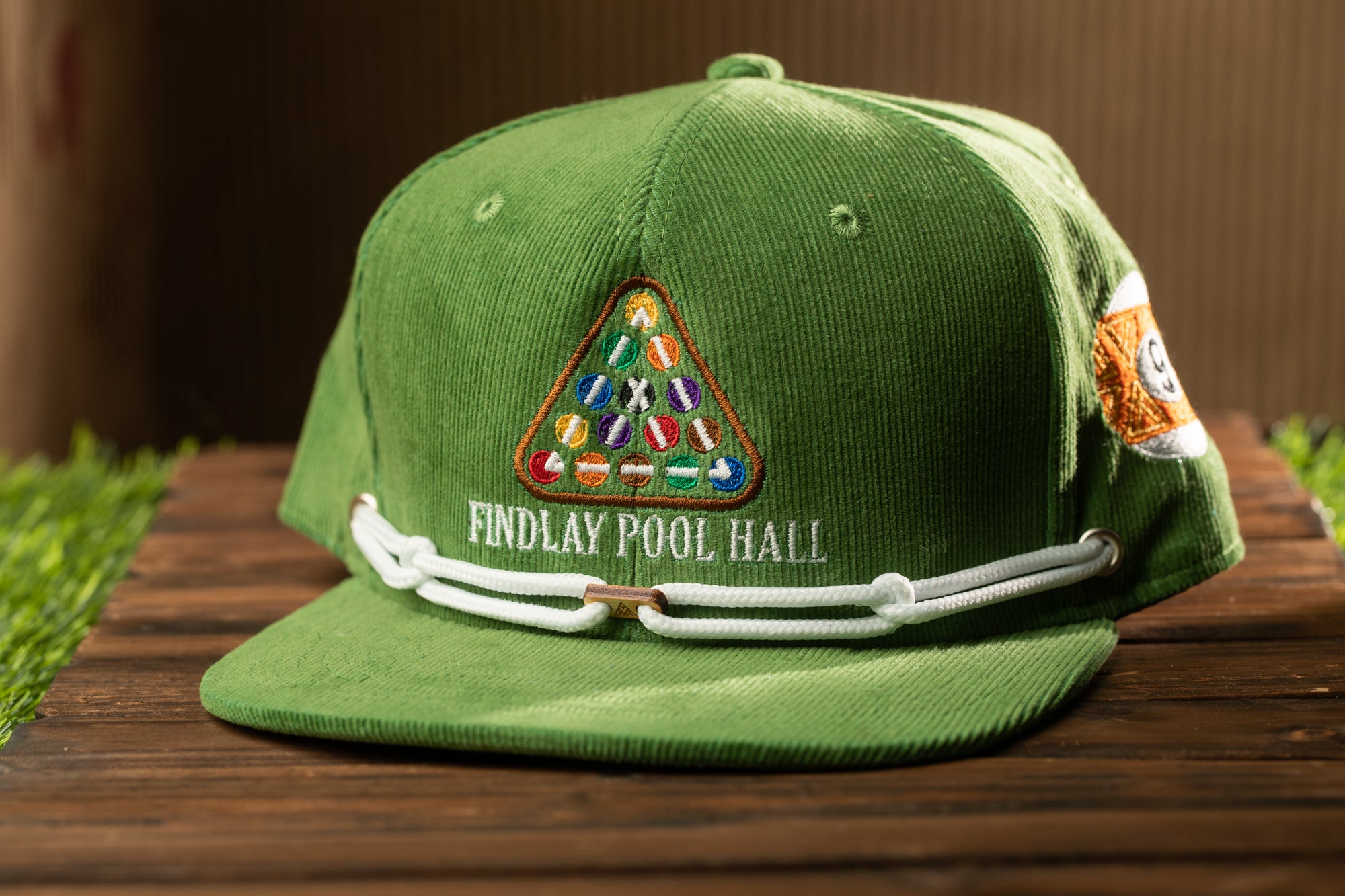A green baseball cap with a pool hall logo and text on it, placed on a wooden surface.
