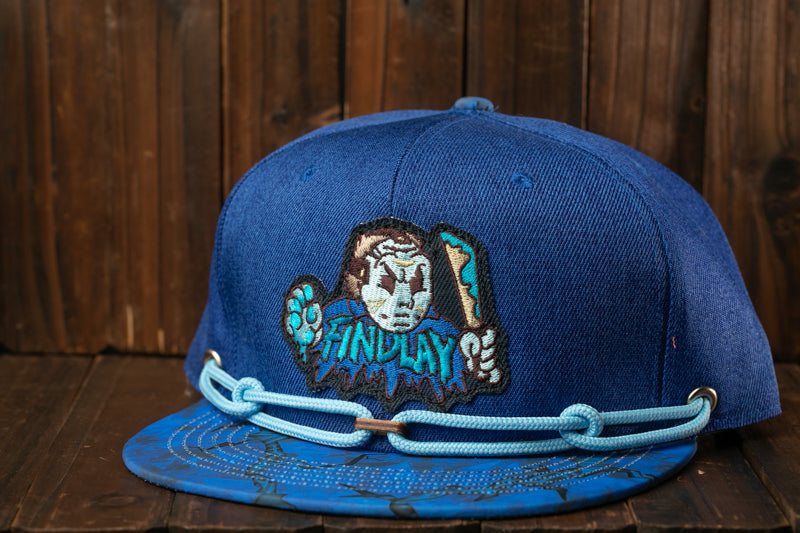 A blue baseball cap with a cartoon character design and the word "FINDPLAY" printed on it, sitting on a wooden surface.