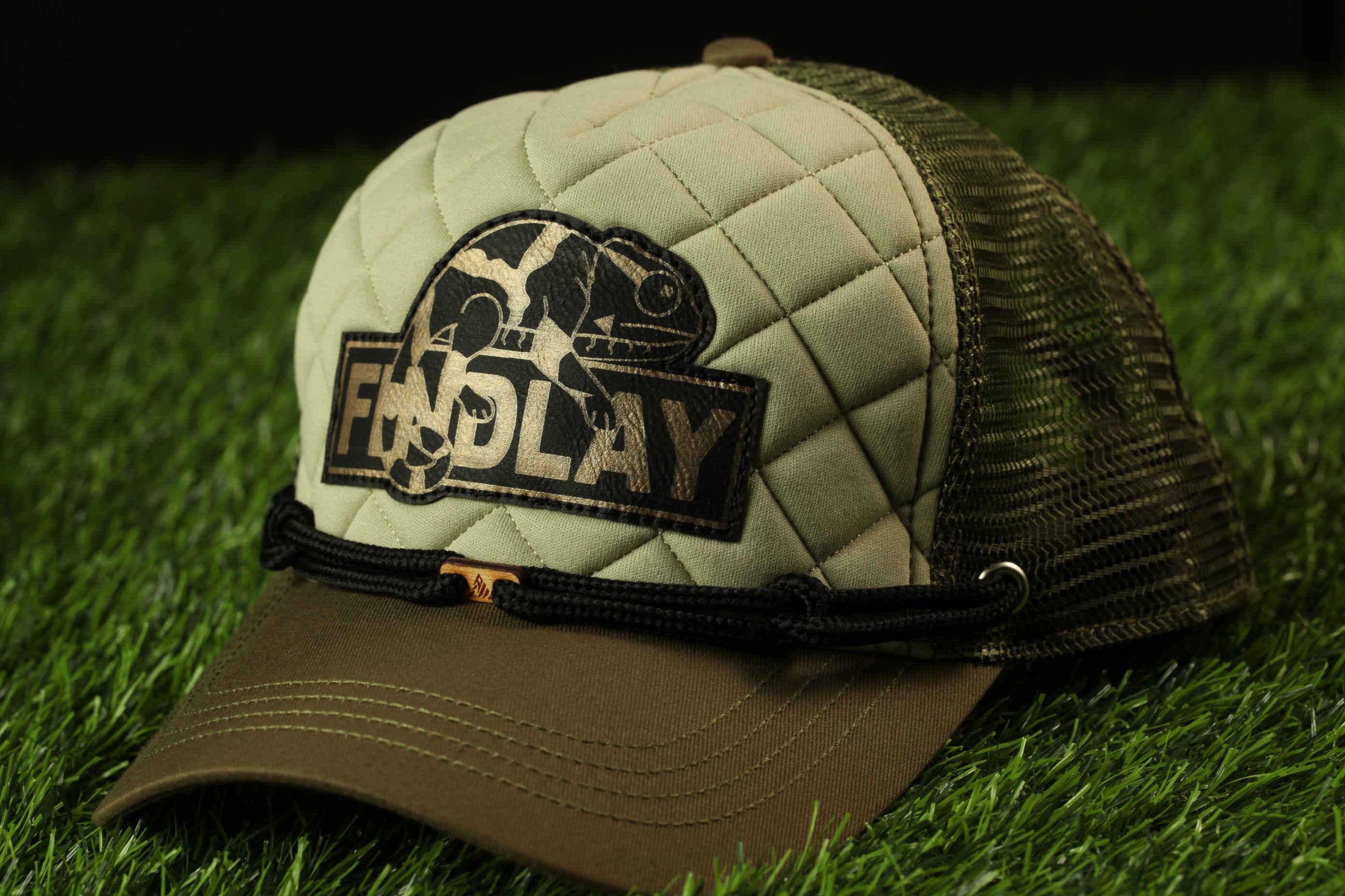 A tan baseball cap with a mesh panel and a black logo on the front, placed on a grassy surface.