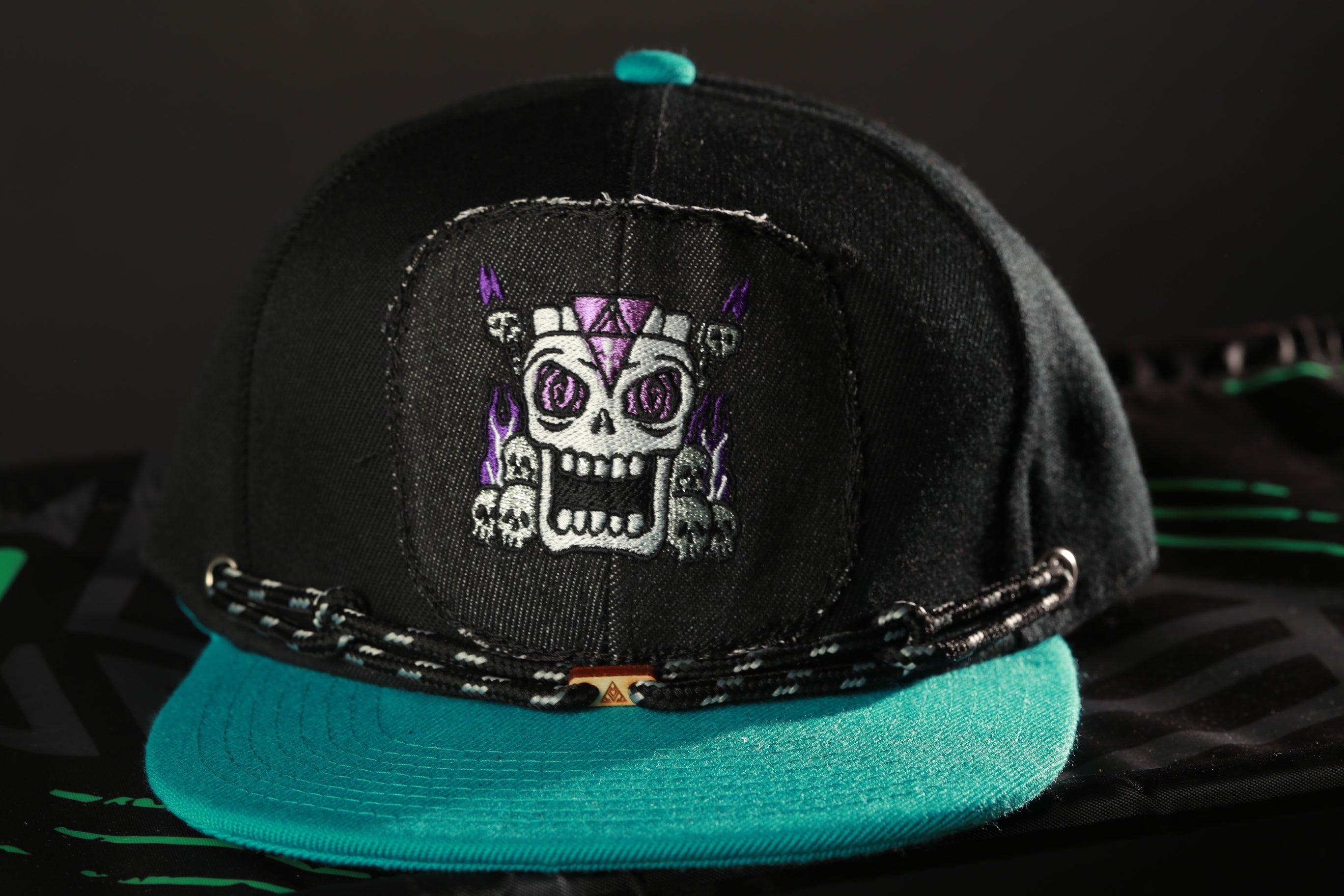 A black baseball cap with a teal brim and a skull design on the front.