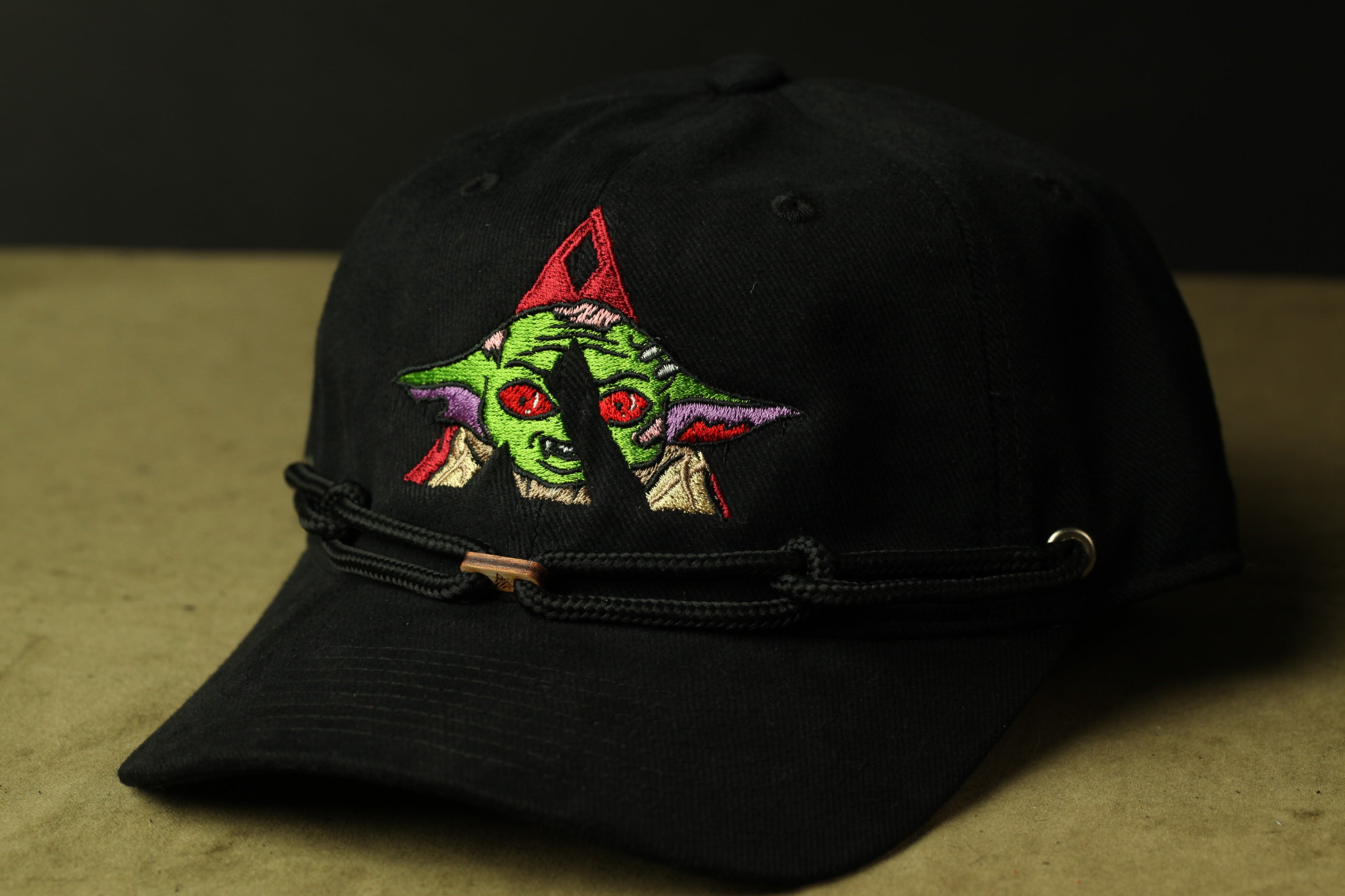 A black baseball cap with a colorful embroidered design of a green alien-like creature on the front.