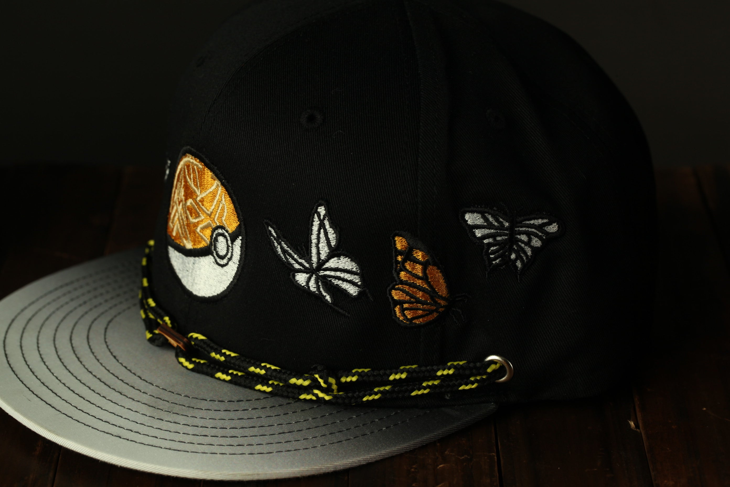 A black baseball cap with a yellow and white butterfly design on the front, sitting on a wooden surface.