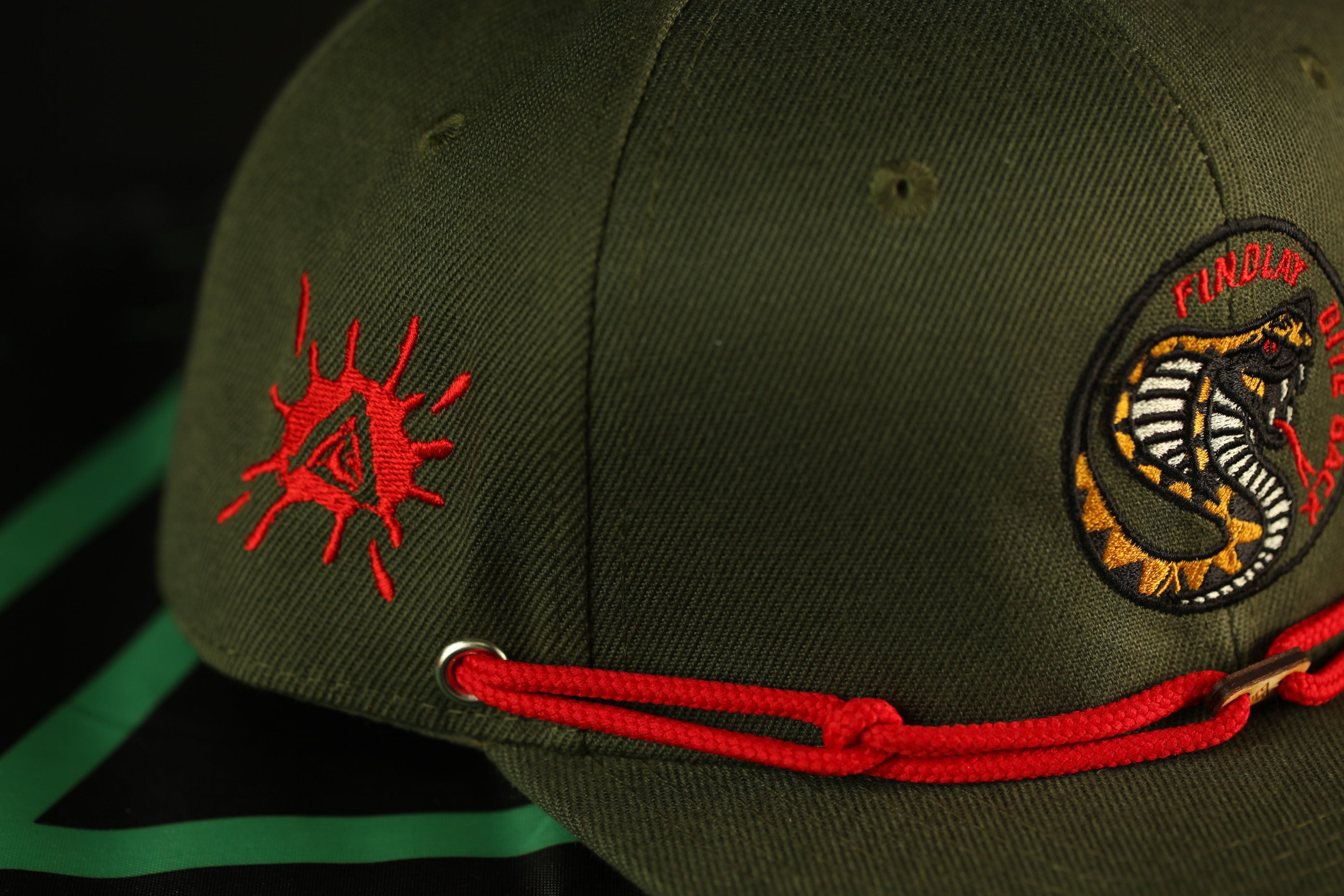 A close-up view of a green baseball cap with red strings and a red and black logo on the front.