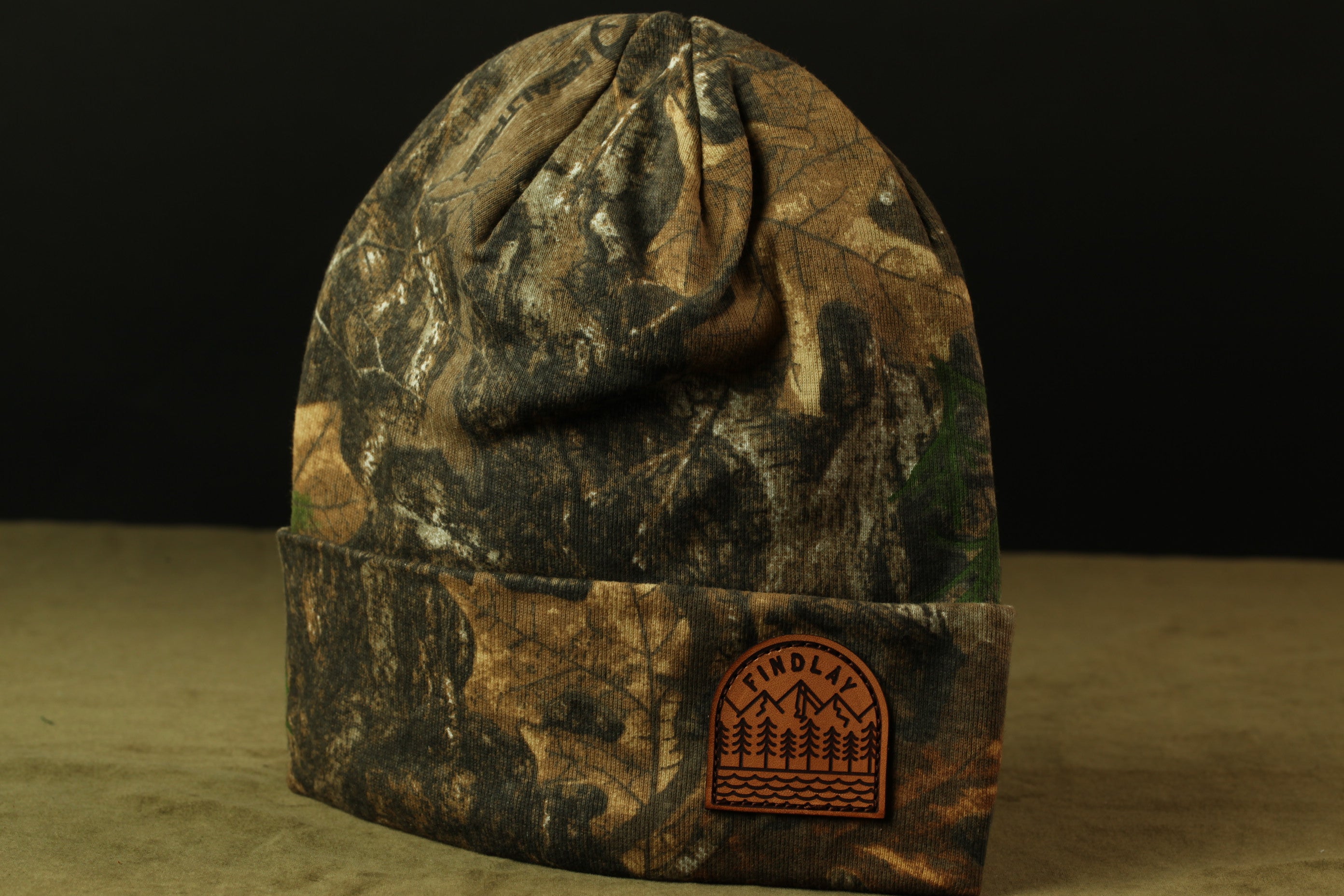A camouflage-patterned beanie with a patch on the front displaying the word "FINOLA".