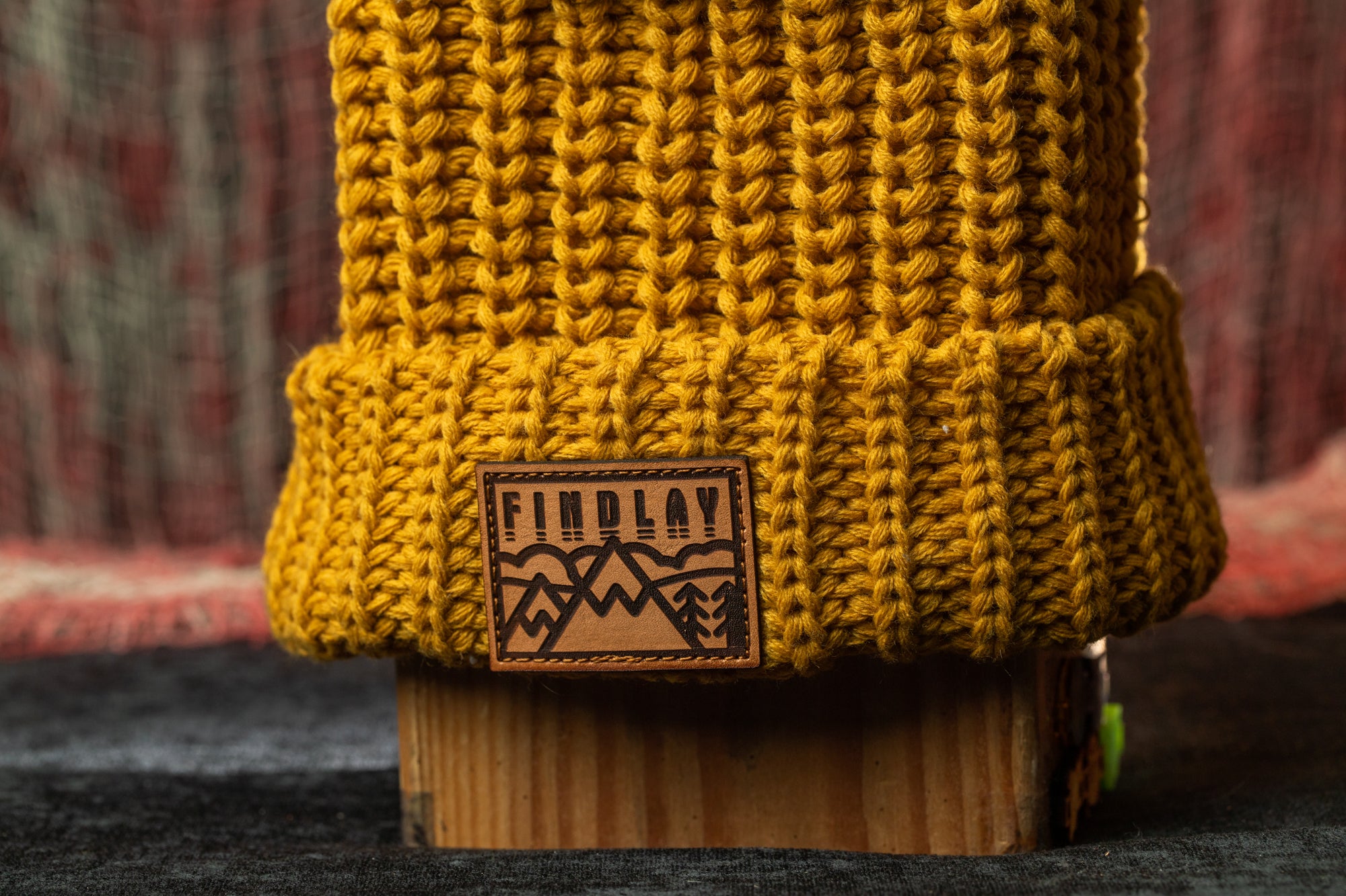 A close-up image of a yellow knitted beanie with a wooden block underneath it, featuring a leather patch with the text "FIND LAY" on it.