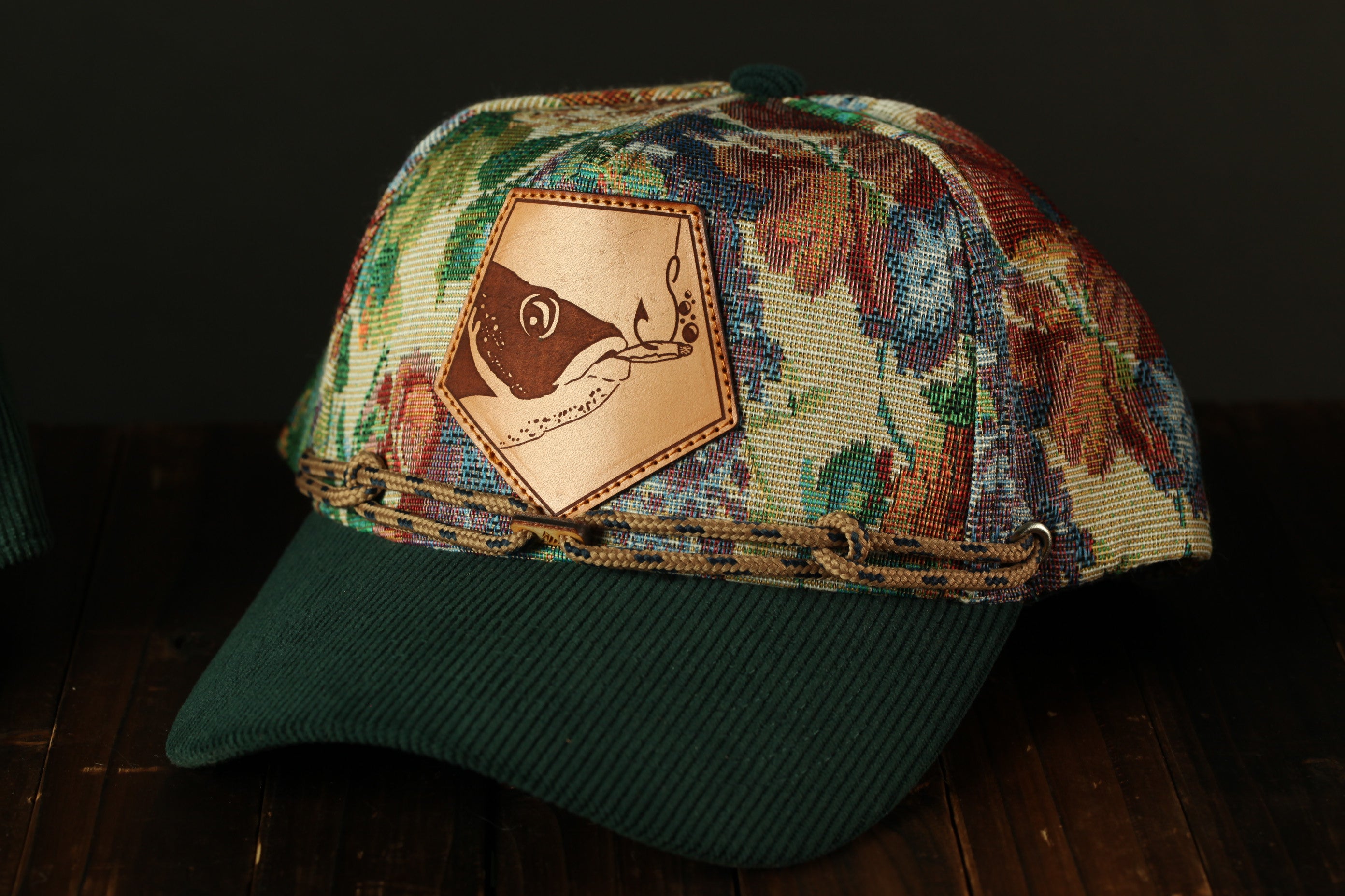 A colorful floral-patterned baseball cap with a leather patch featuring a fish design, placed on a wooden surface.