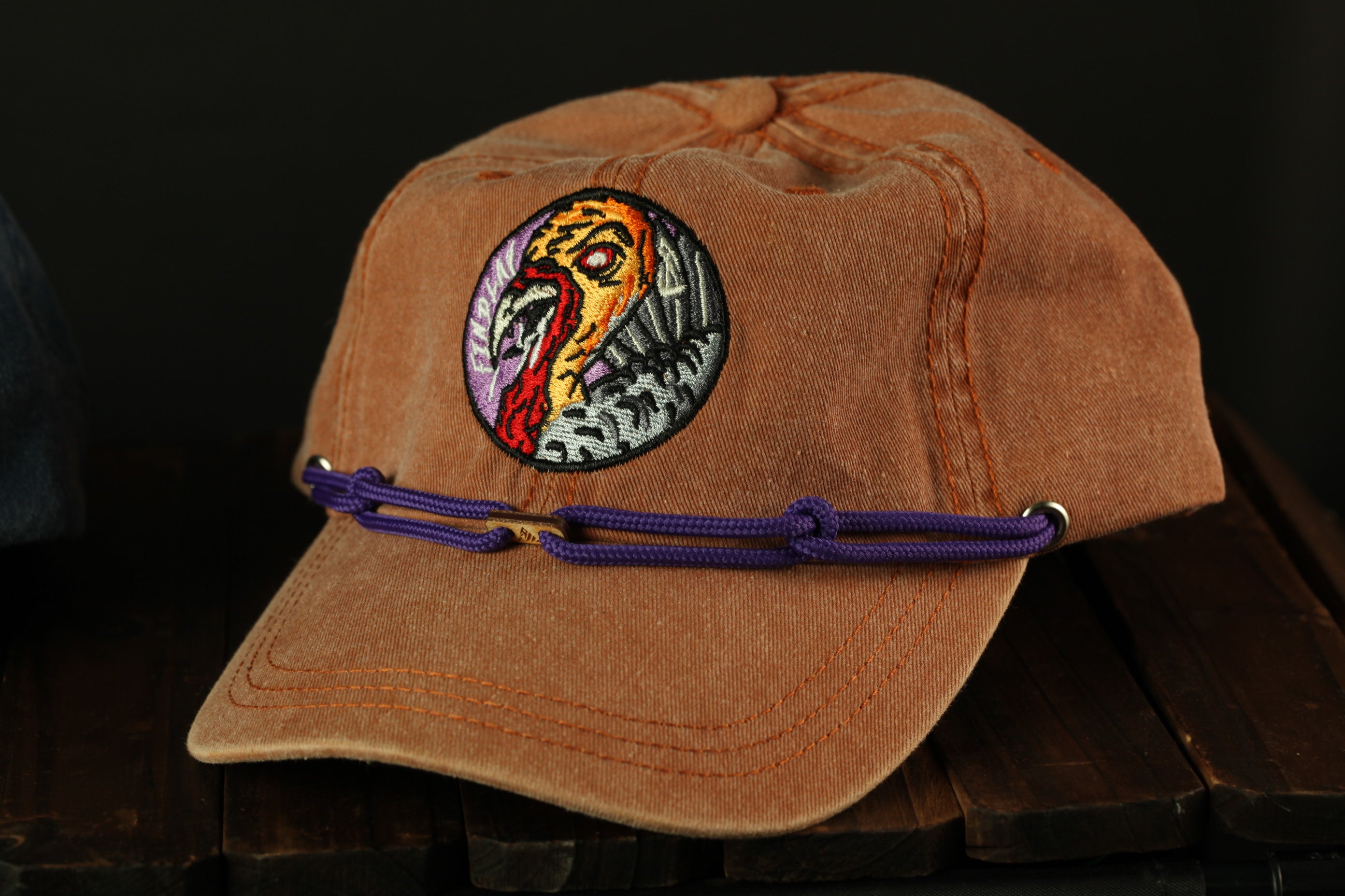 A brown baseball cap with a colorful embroidered design on the front, featuring a bird-like motif.