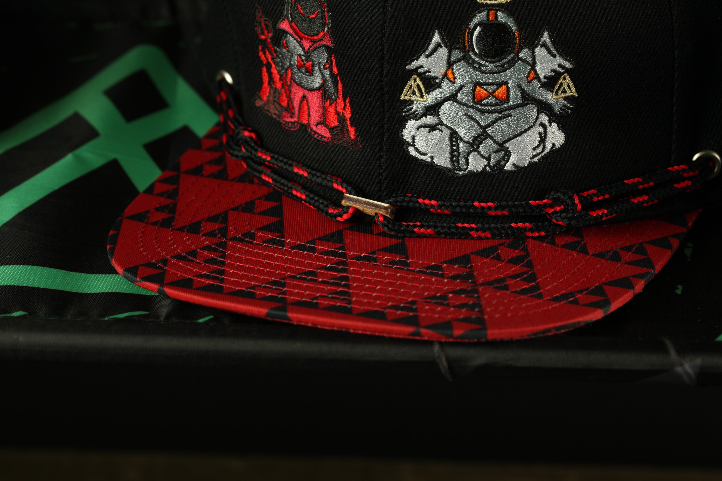 A close-up view of a black baseball cap with a red and black geometric pattern on the brim and a white astronaut figure on the front.