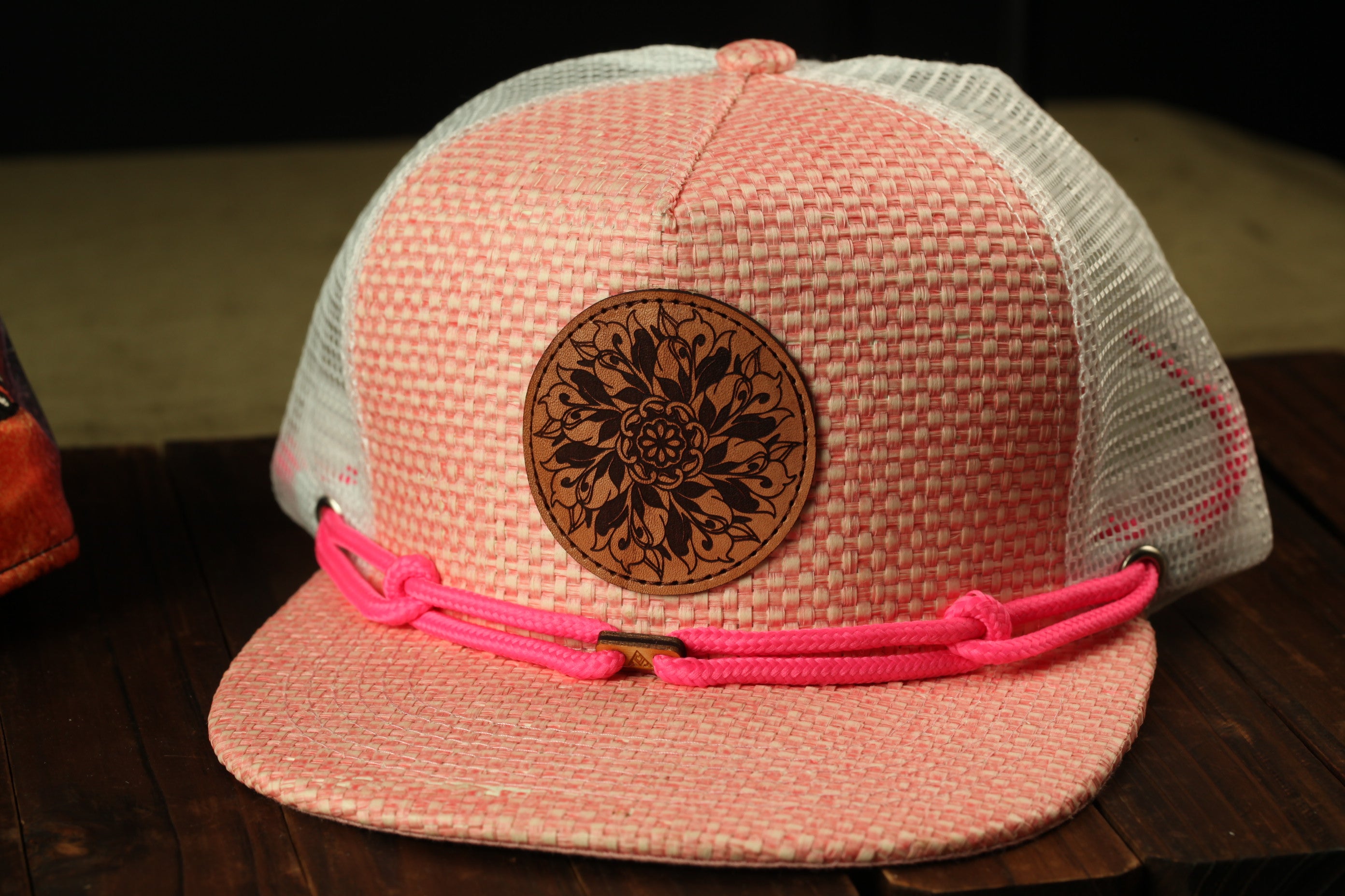 A pink and white patterned baseball cap with a circular leather patch and pink braided straps is displayed on a wooden surface.