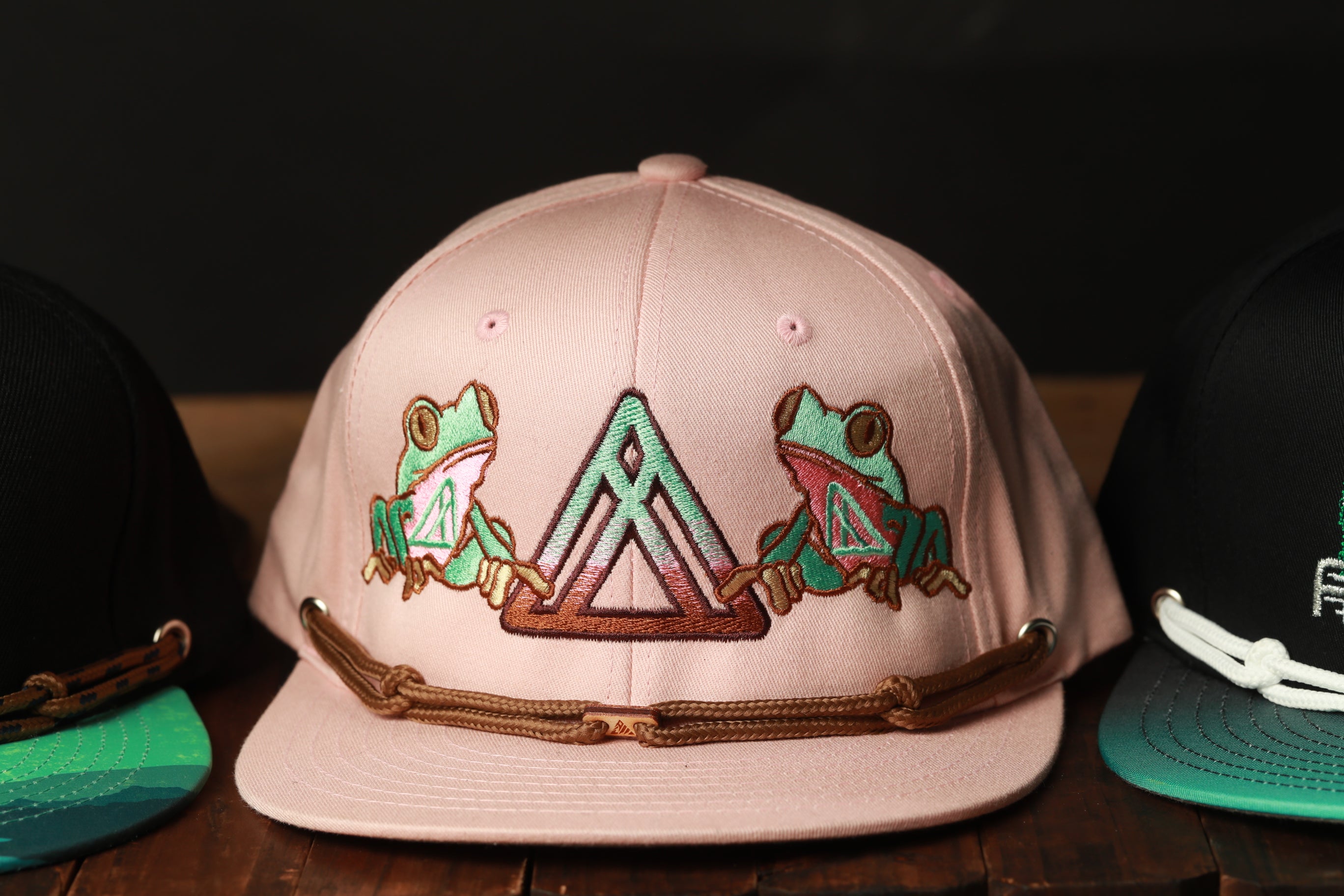 A pink baseball cap with a graphic of two green frogs on it, sitting on a wooden surface.