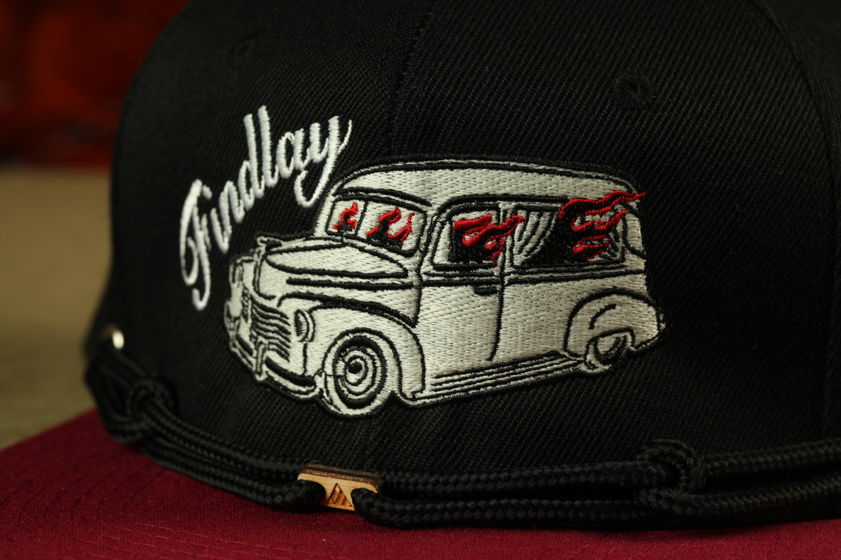 A close-up view of a black baseball cap with a white and red embroidered design depicting a classic car.