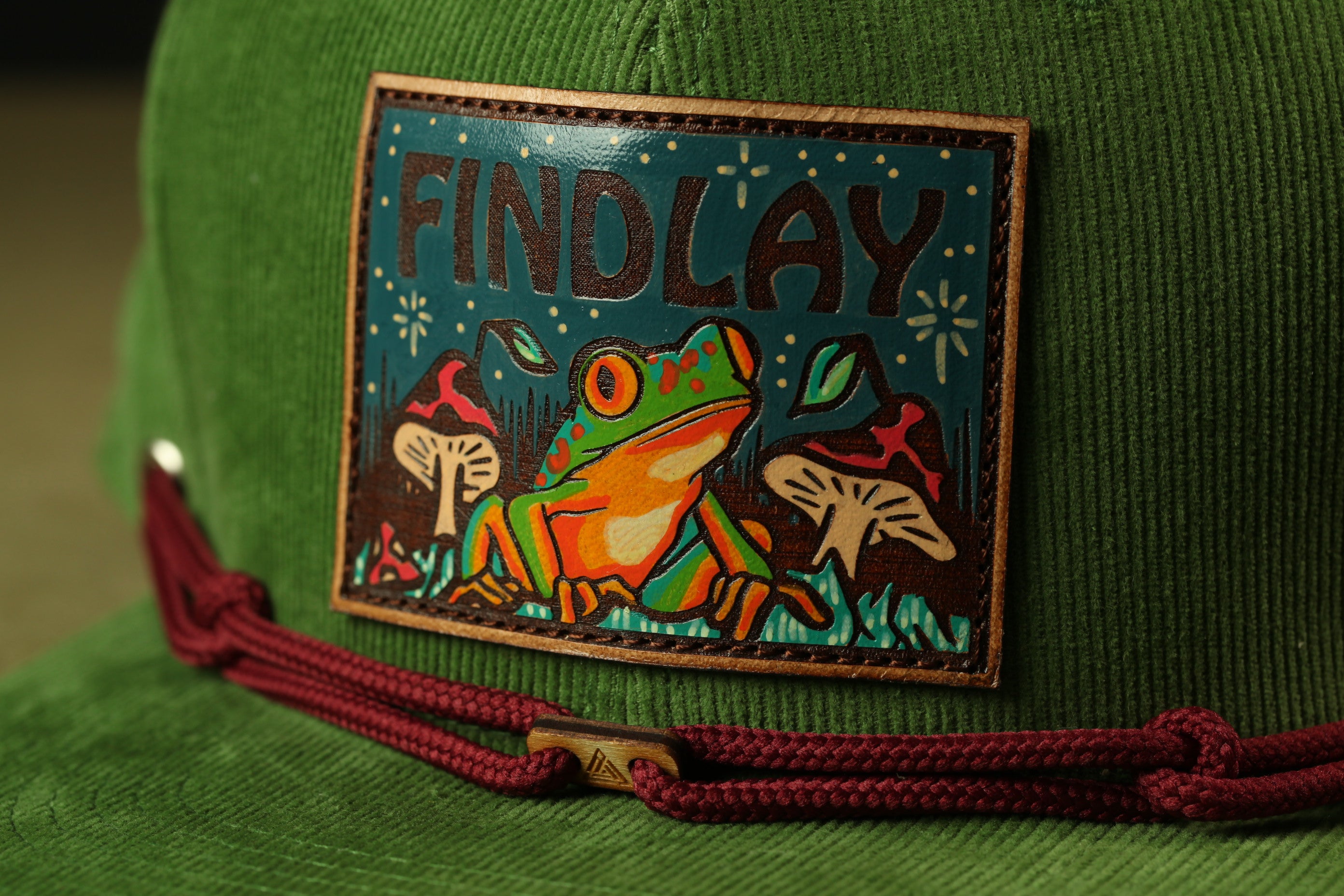 A close-up of a green hat with a patch featuring a frog and mushrooms, along with the word "FINDLAY" printed on it.