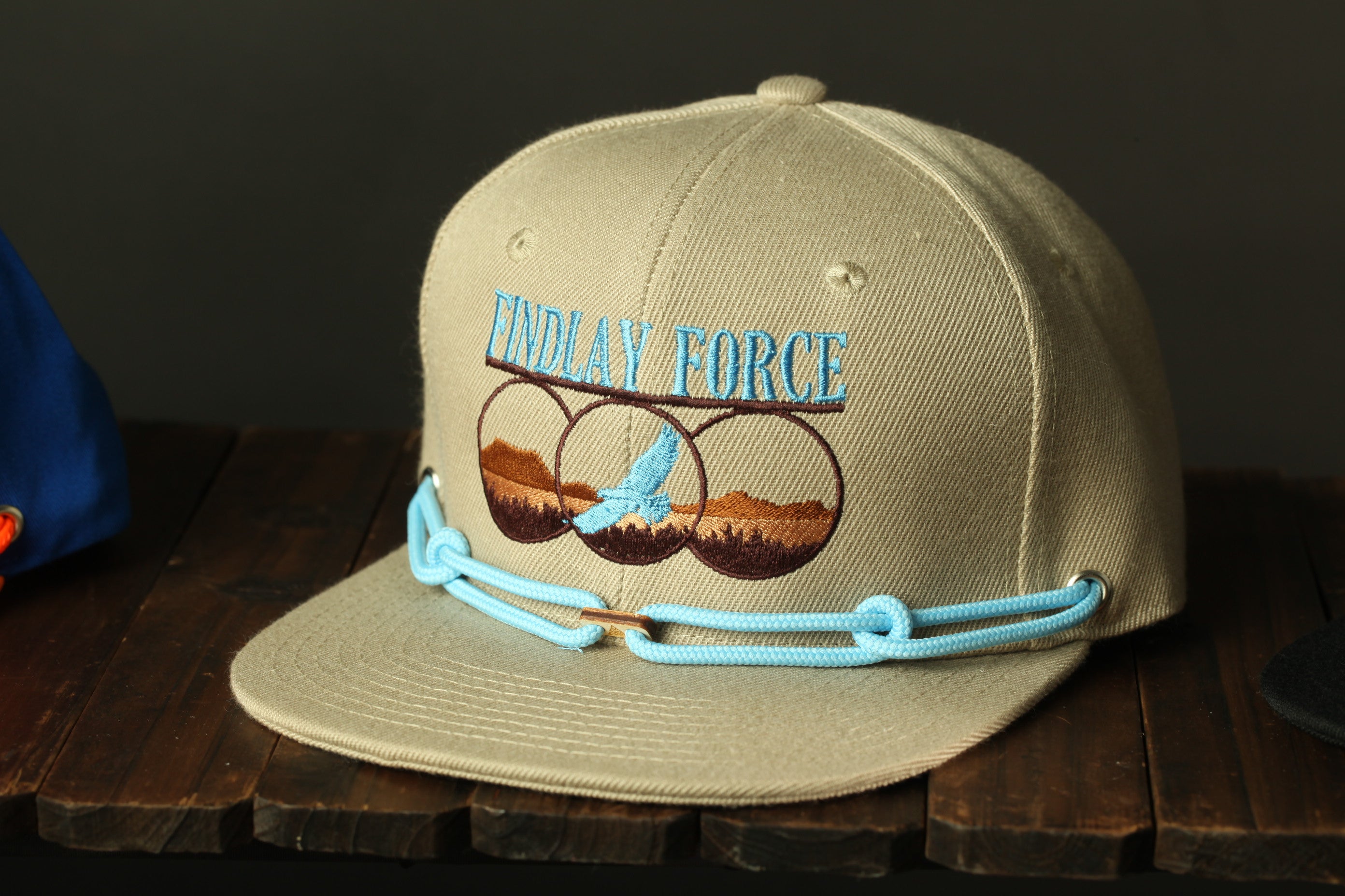 A tan baseball cap with a blue and brown embroidered design, featuring a bird and mountains, sits on a wooden surface.