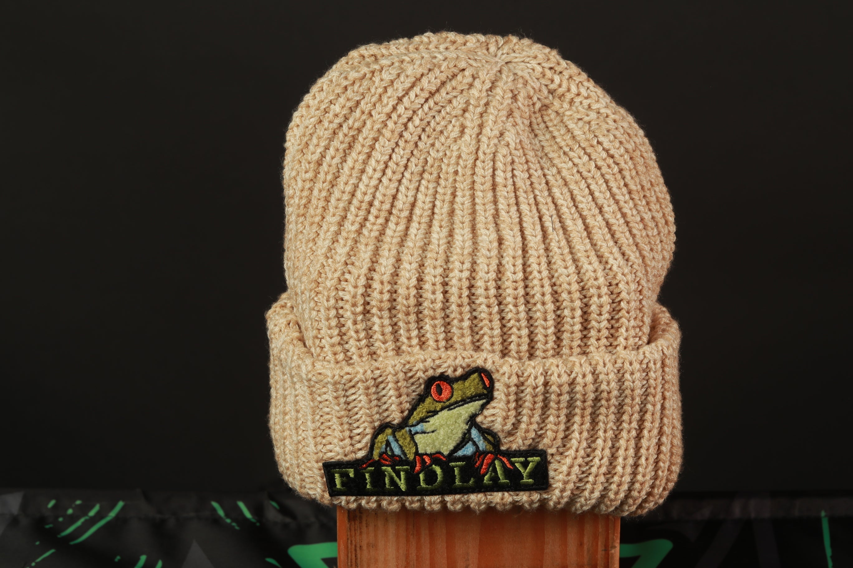 A beige knit beanie with a frog embroidered on it, placed on a wooden surface against a black background.