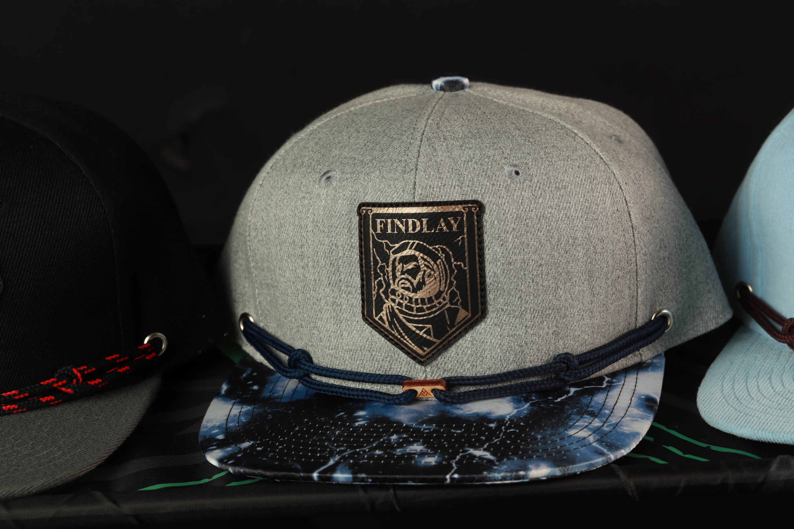 A gray baseball cap with a black and white tie-dye pattern and a "Findlay" patch on the front, sitting on a shelf among other hats.