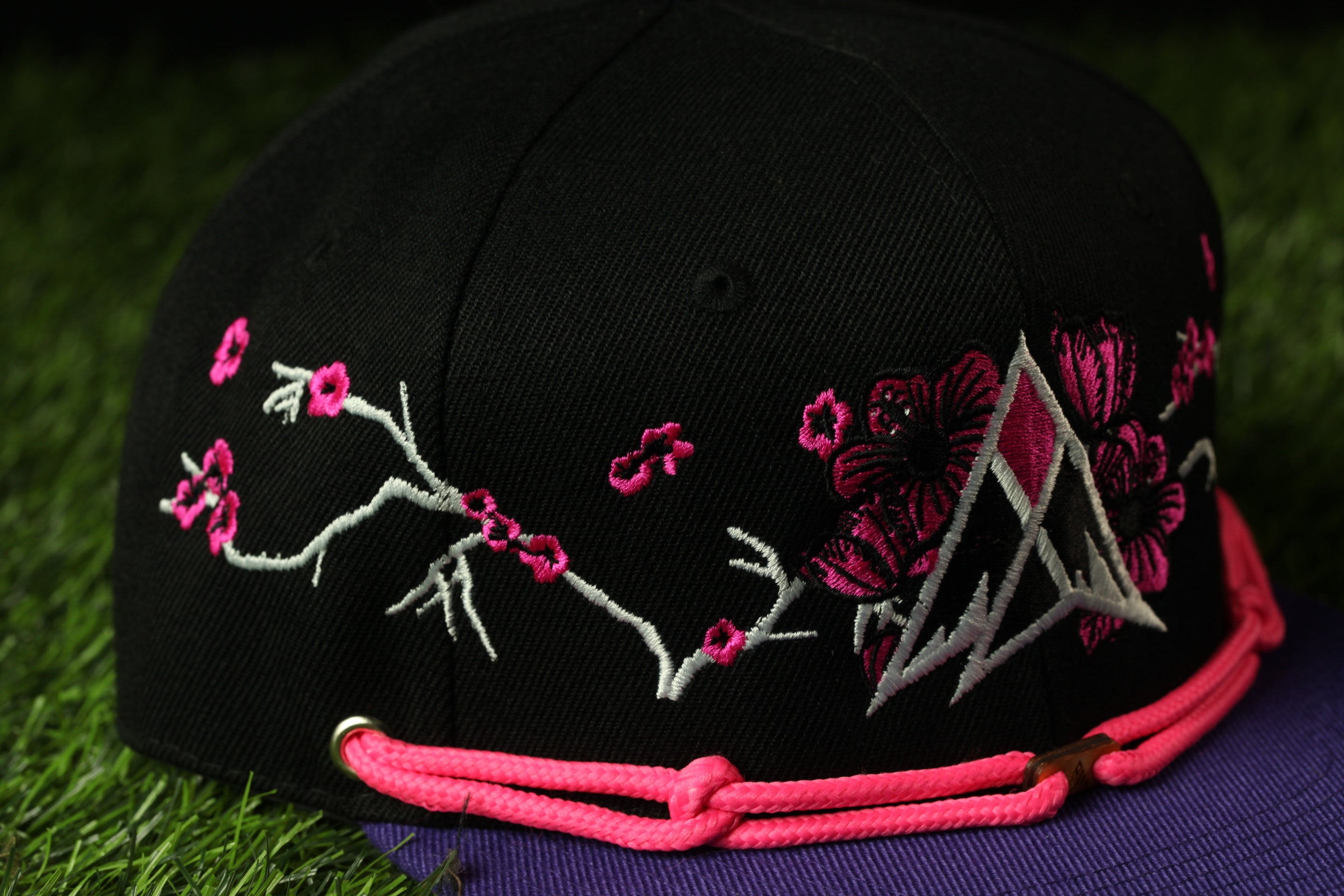 A black baseball cap with pink floral embroidery and a pink braided strap is shown against a green background.