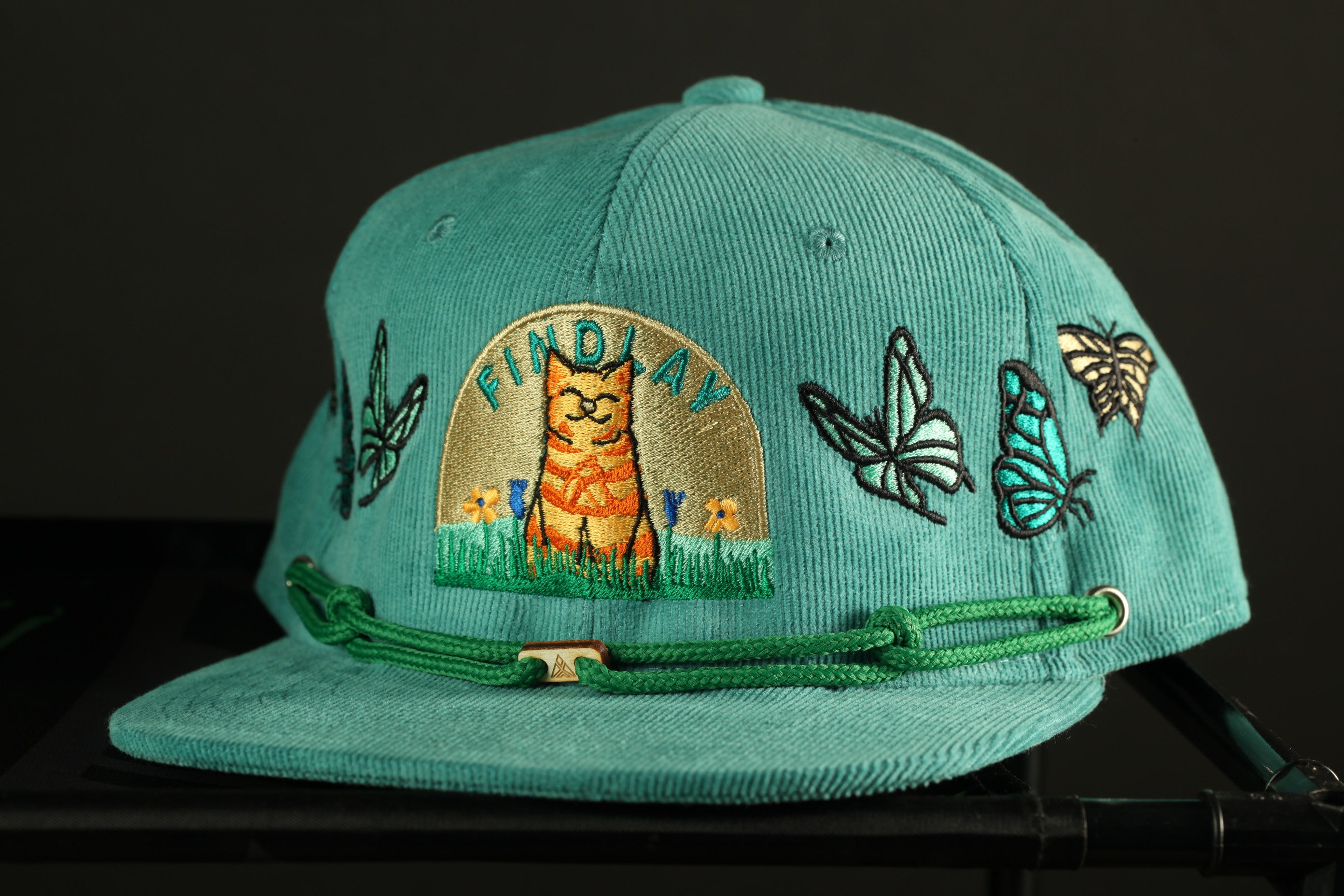 A turquoise baseball cap with various embroidered designs, including a cat, butterflies, and flowers, as well as the text "FIND A WAY" on the front.