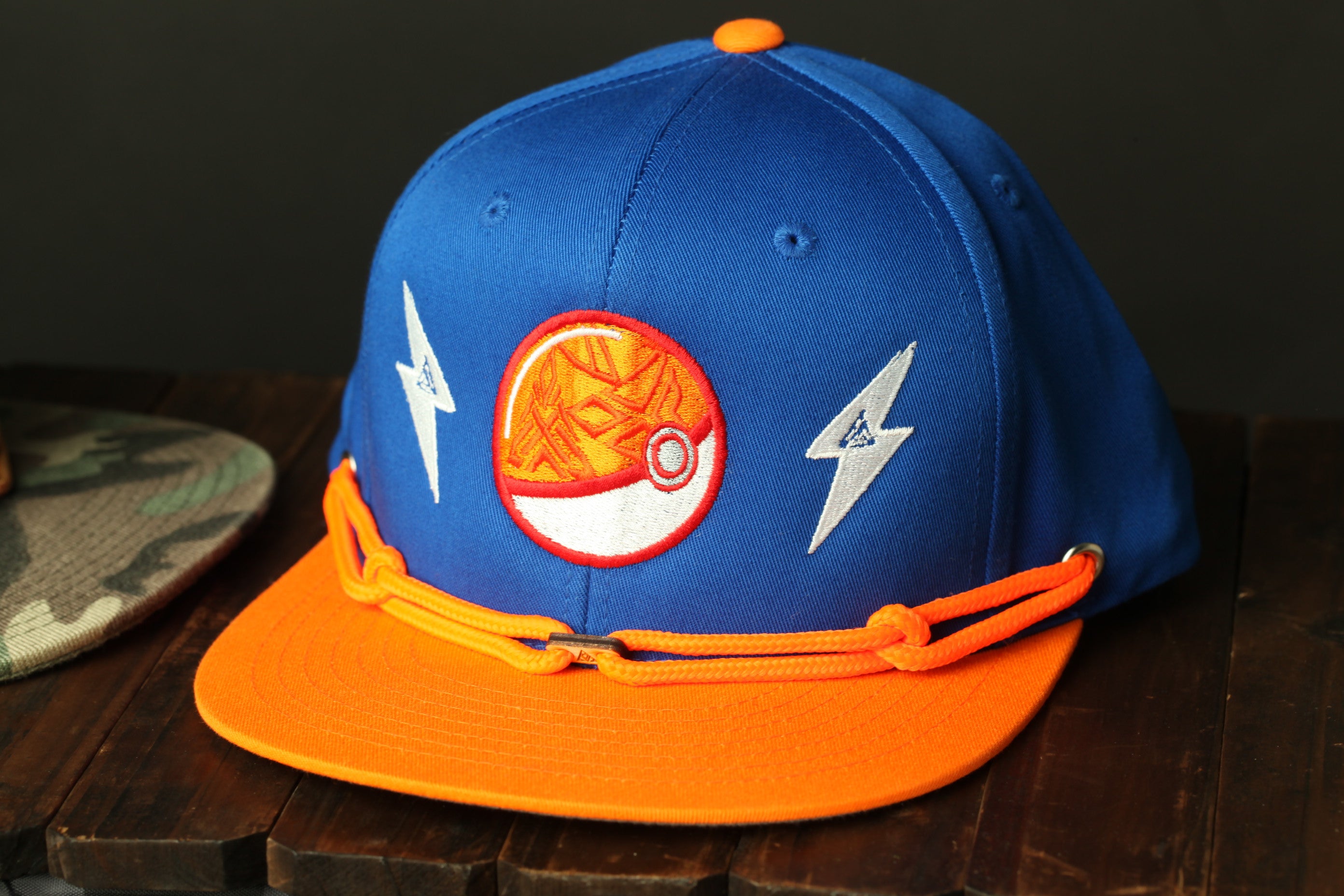 A blue baseball cap with orange accents and a Pokemon-themed patch is displayed on a wooden surface.