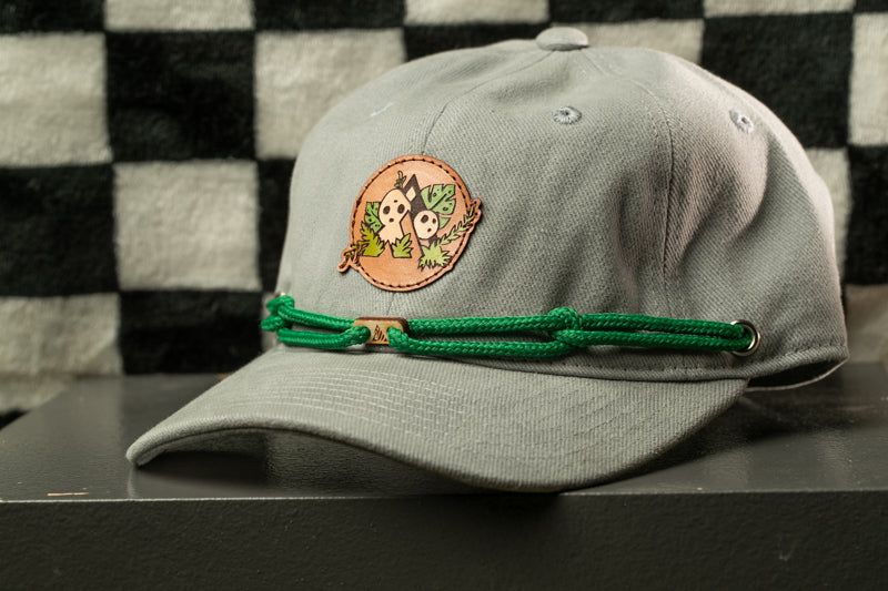 A gray baseball cap with a green string and a circular patch featuring cartoon characters is displayed against a black and white checkered background.