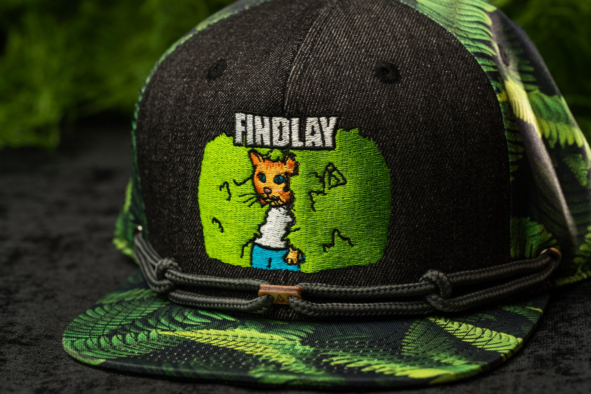 A black baseball cap with a green and black camouflage pattern, featuring a cartoon image of a cat and the word "FINDlay" printed on it.