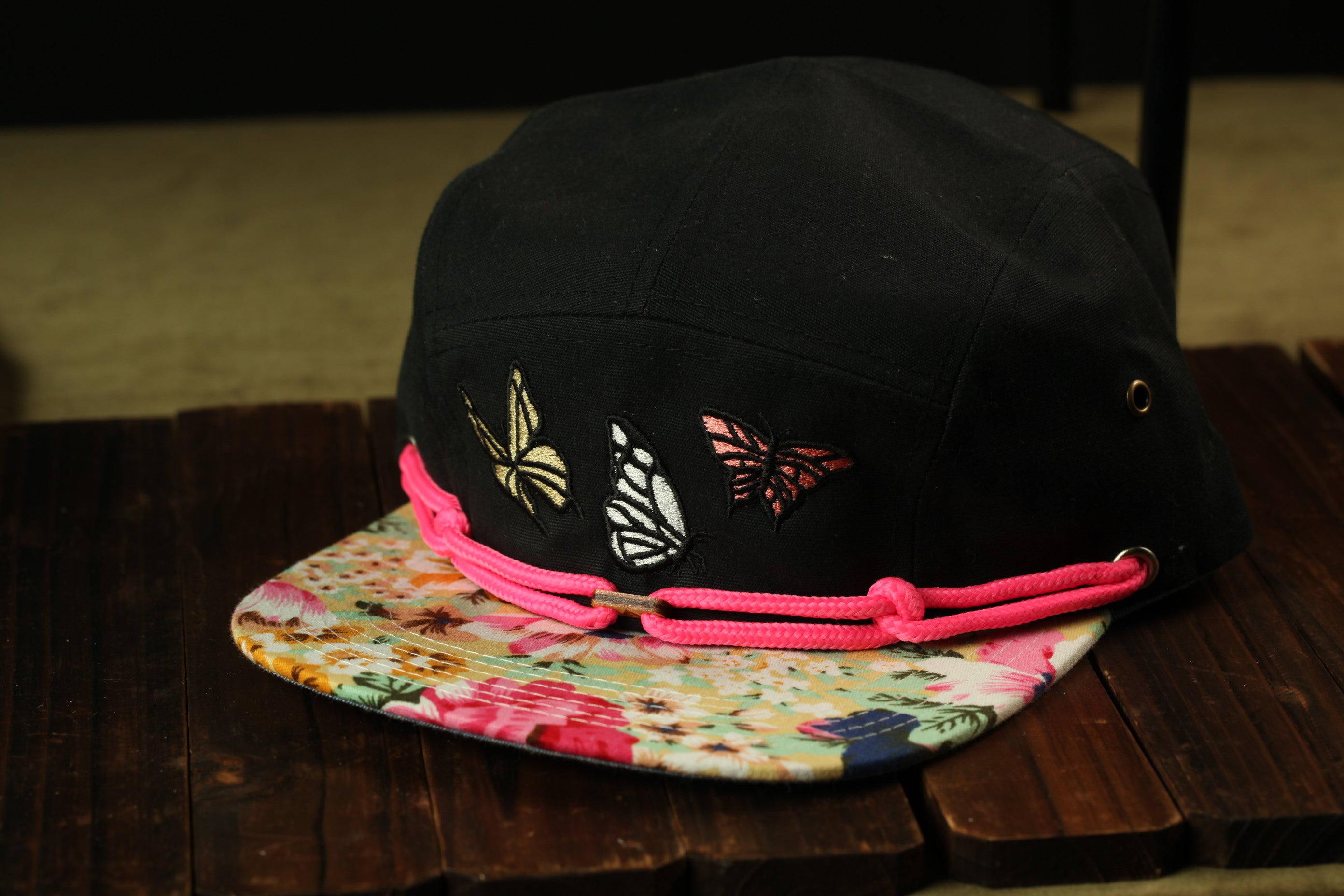 A black baseball cap with colorful butterfly embroidery and a floral pattern on the brim, resting on a wooden surface.