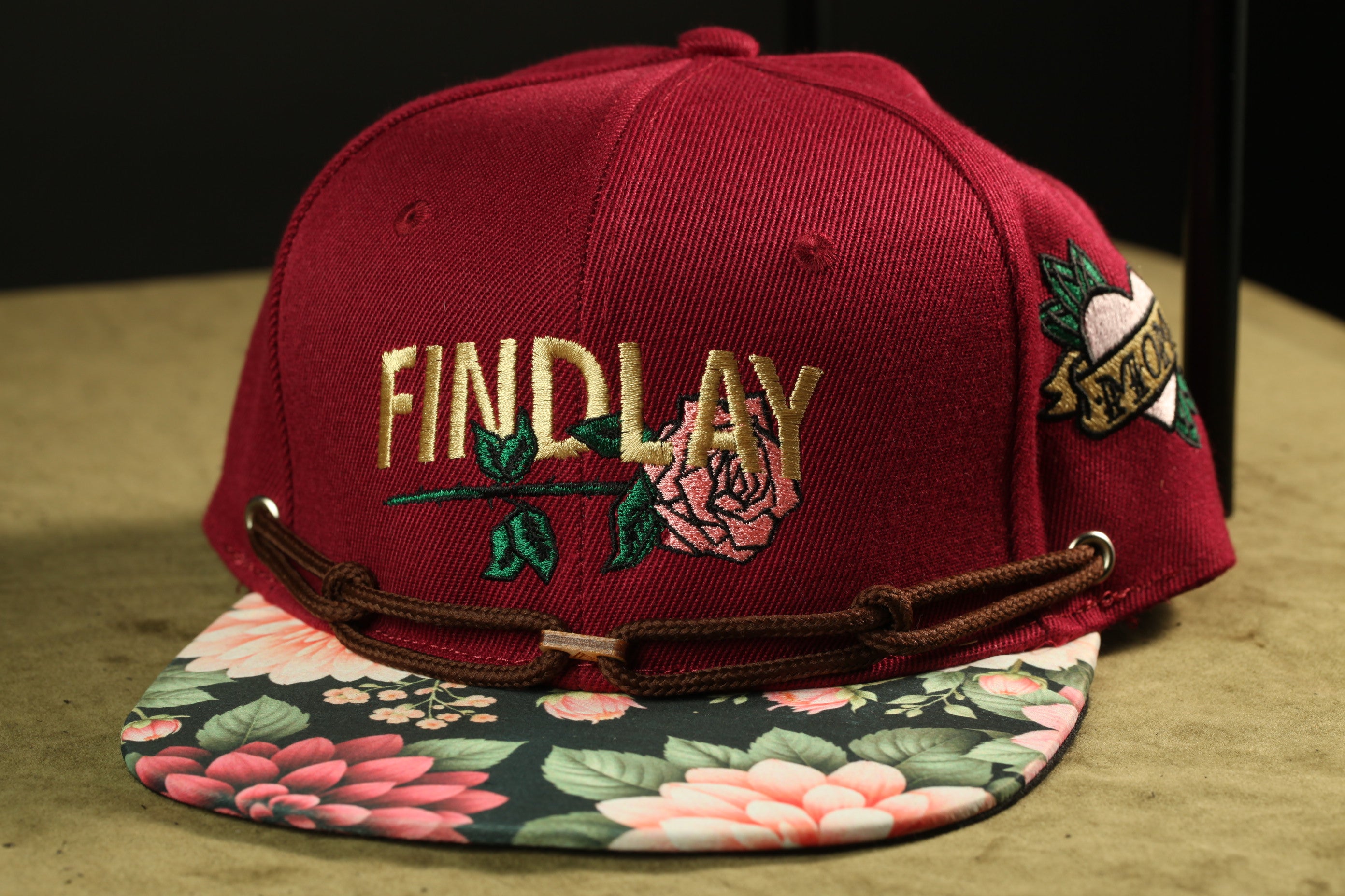A maroon baseball cap with the word "FINDlay" embroidered on it, featuring a floral pattern on the brim and a chain strap.