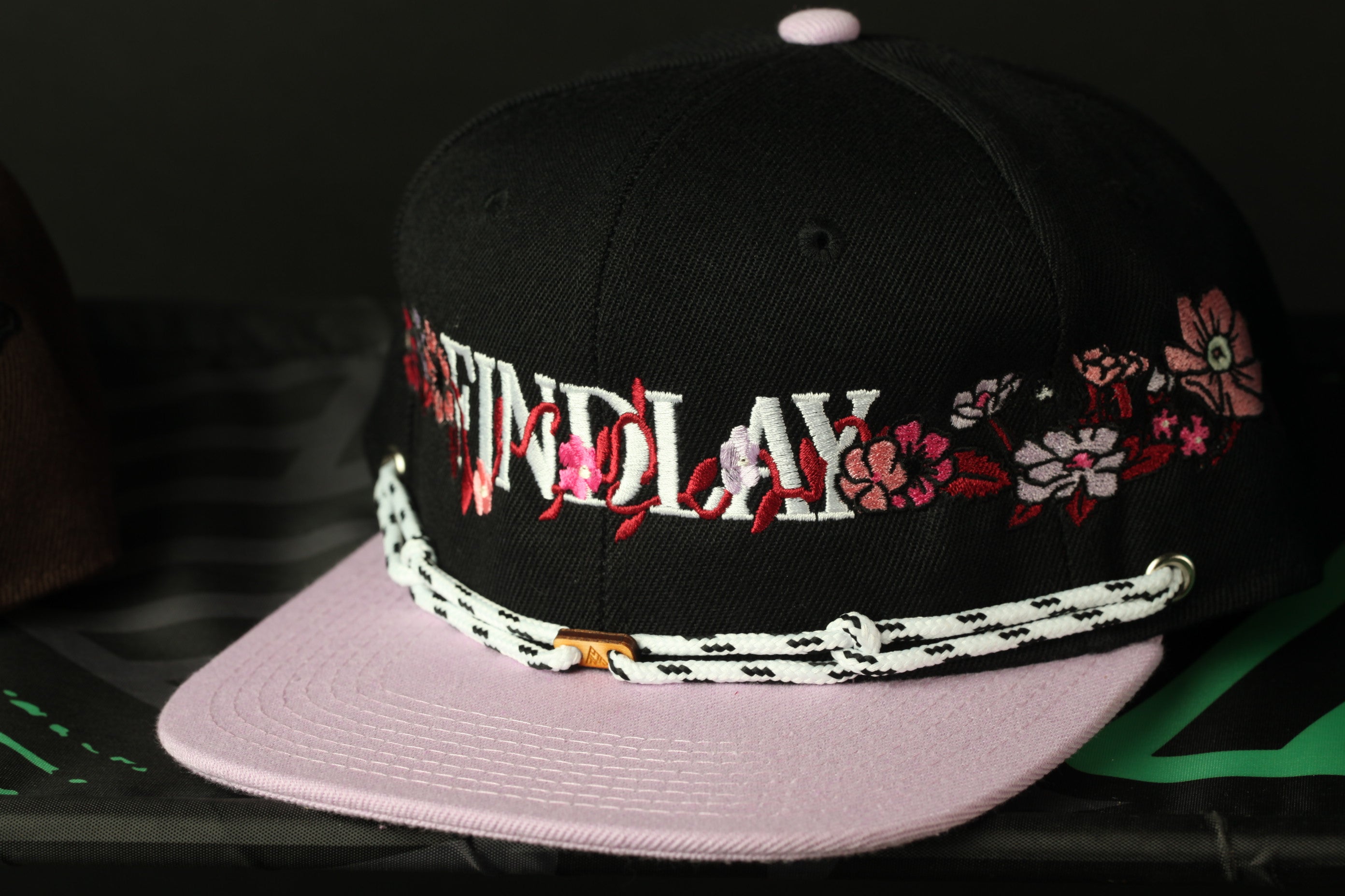 A black baseball cap with pink floral embroidery and a white lace strap.