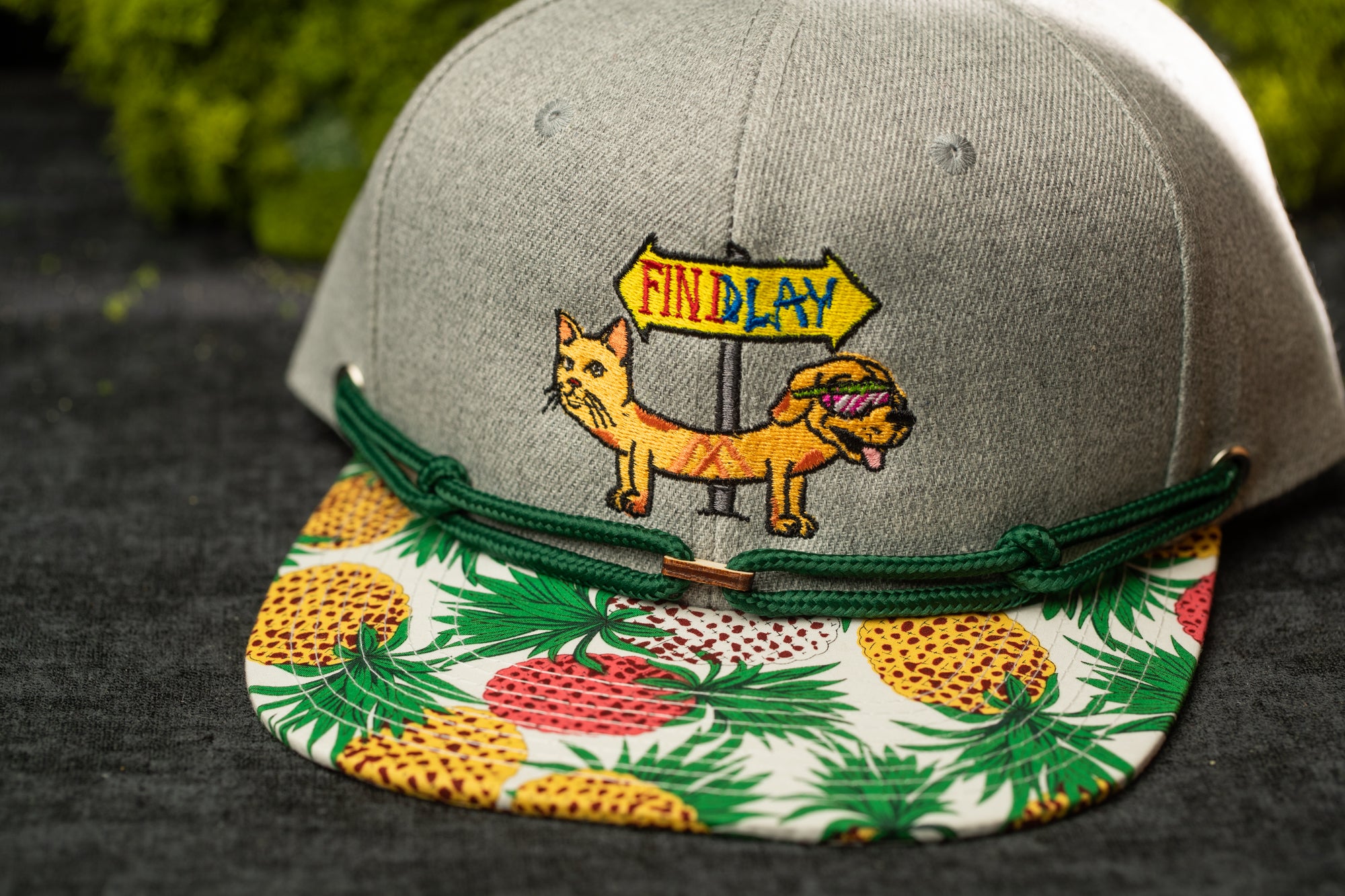 A gray baseball cap with a colorful pineapple pattern and a cartoon cat wearing sunglasses.