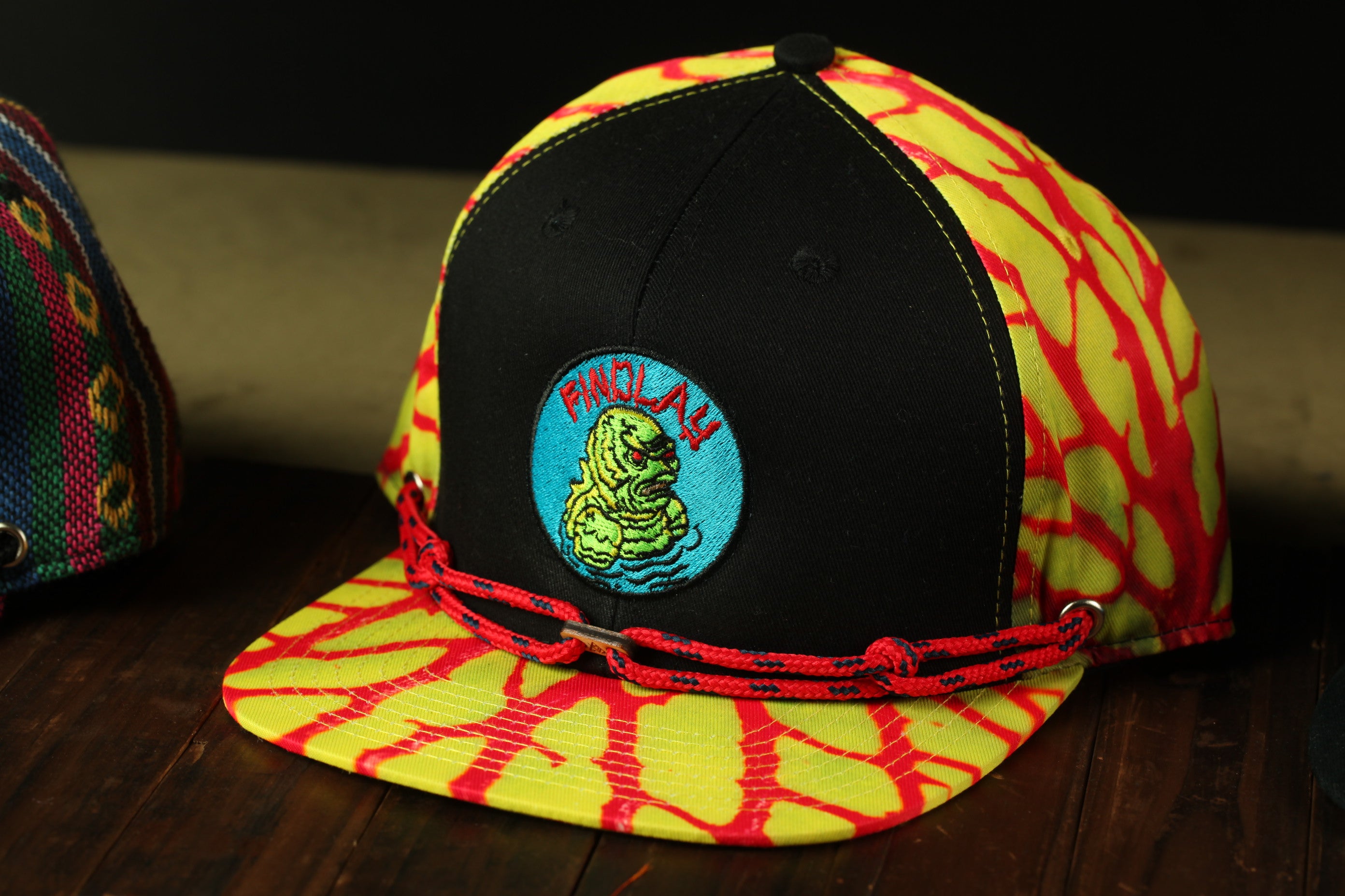 A colorful baseball cap with a black brim and a yellow and red patterned fabric. The cap has a circular patch on the front with a cartoon character and the word "PANOPLAY" written on it.