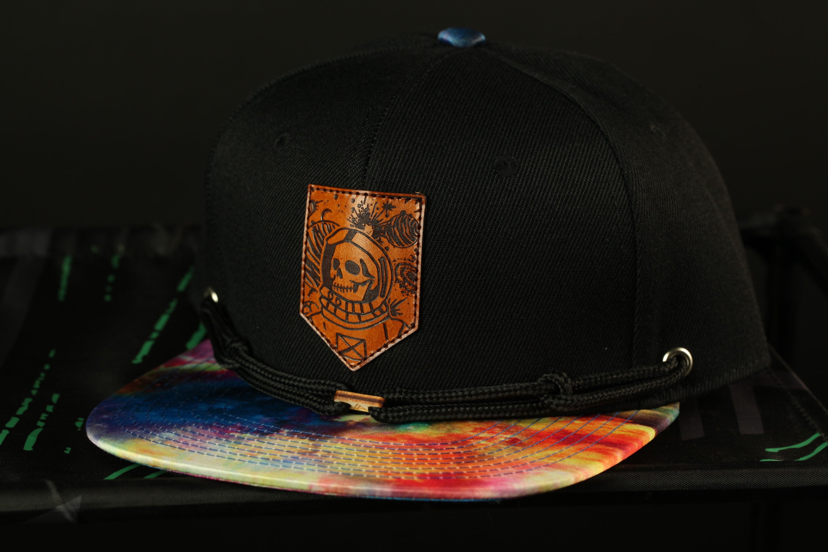 A black baseball cap with a colorful tie-dye pattern and a skull-like patch on the front, placed on a colorful patterned surface.