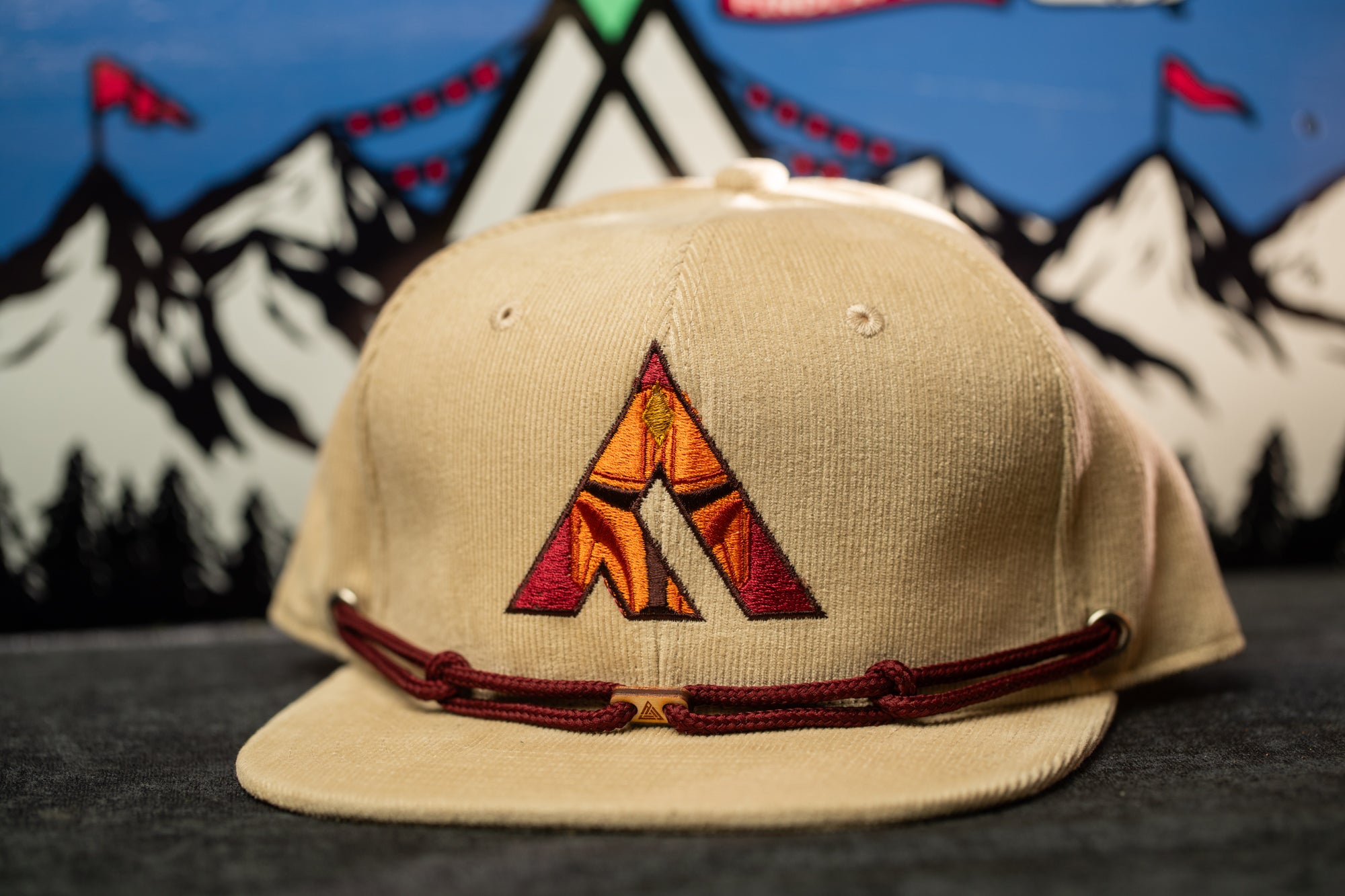A beige baseball cap with a red and orange embroidered logo on the front, placed on a gray surface against a background of mountains and flags.