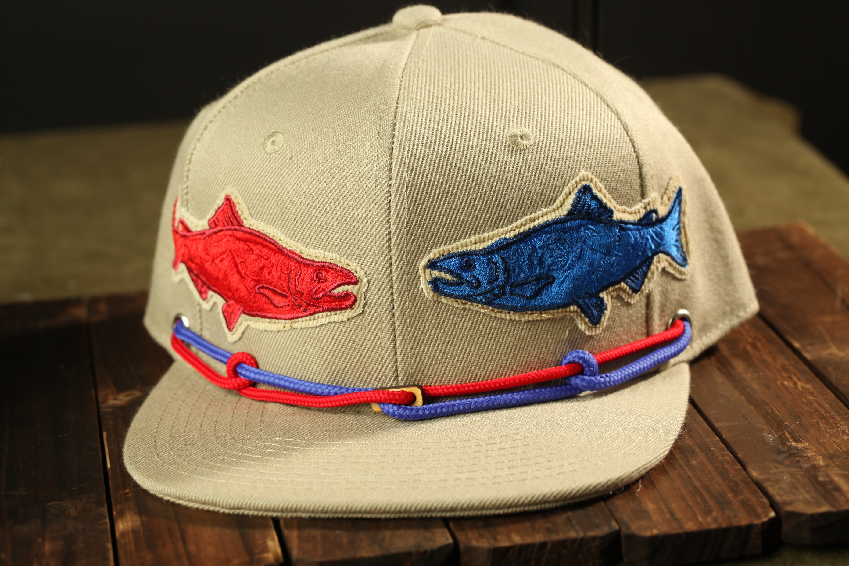 A beige baseball cap with two fish embroidered on it, one red and one blue, and a red and blue string or strap around the brim.