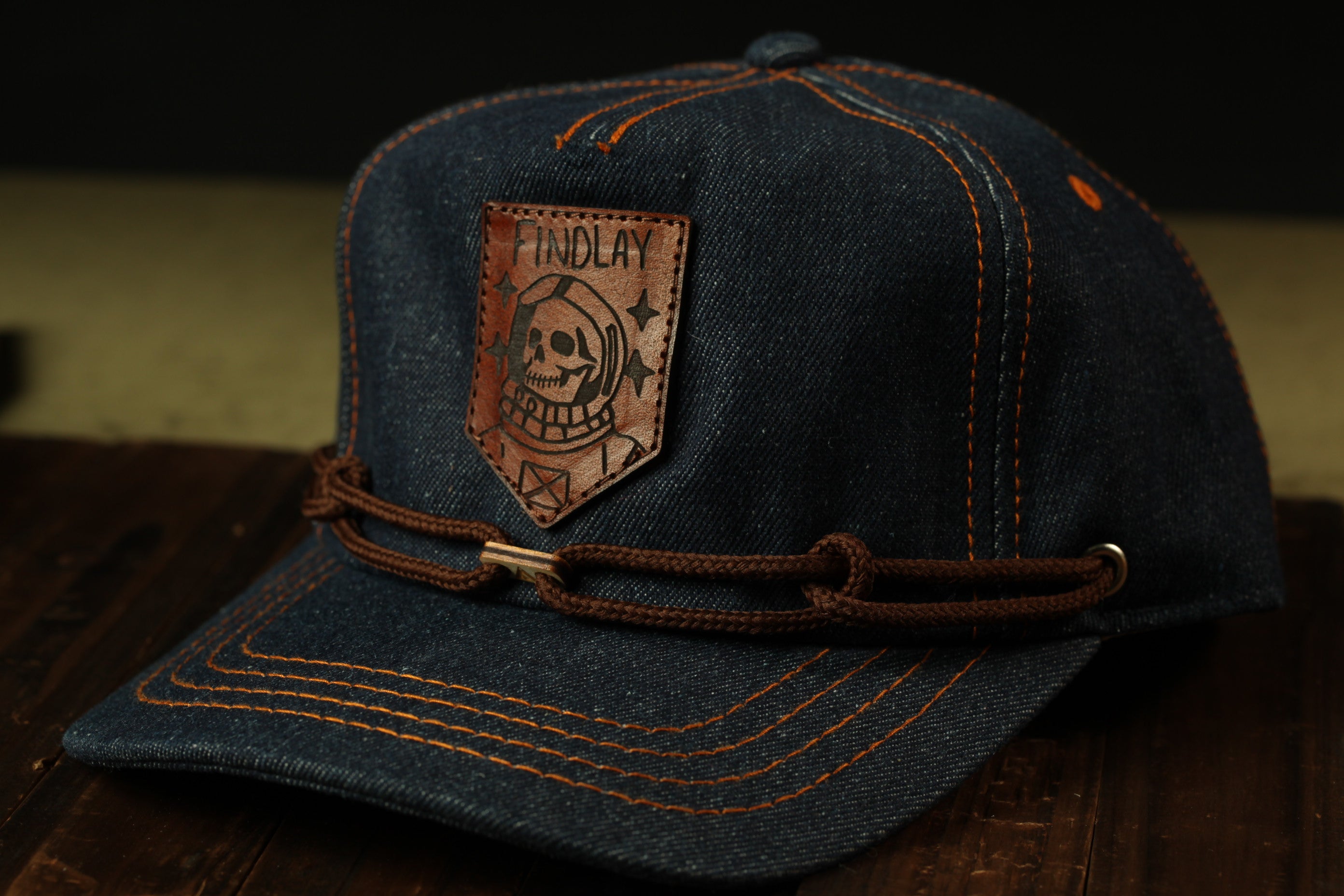 A dark blue denim baseball cap with a brown leather patch featuring a skull and crossbones design, and a brown leather strap across the front.