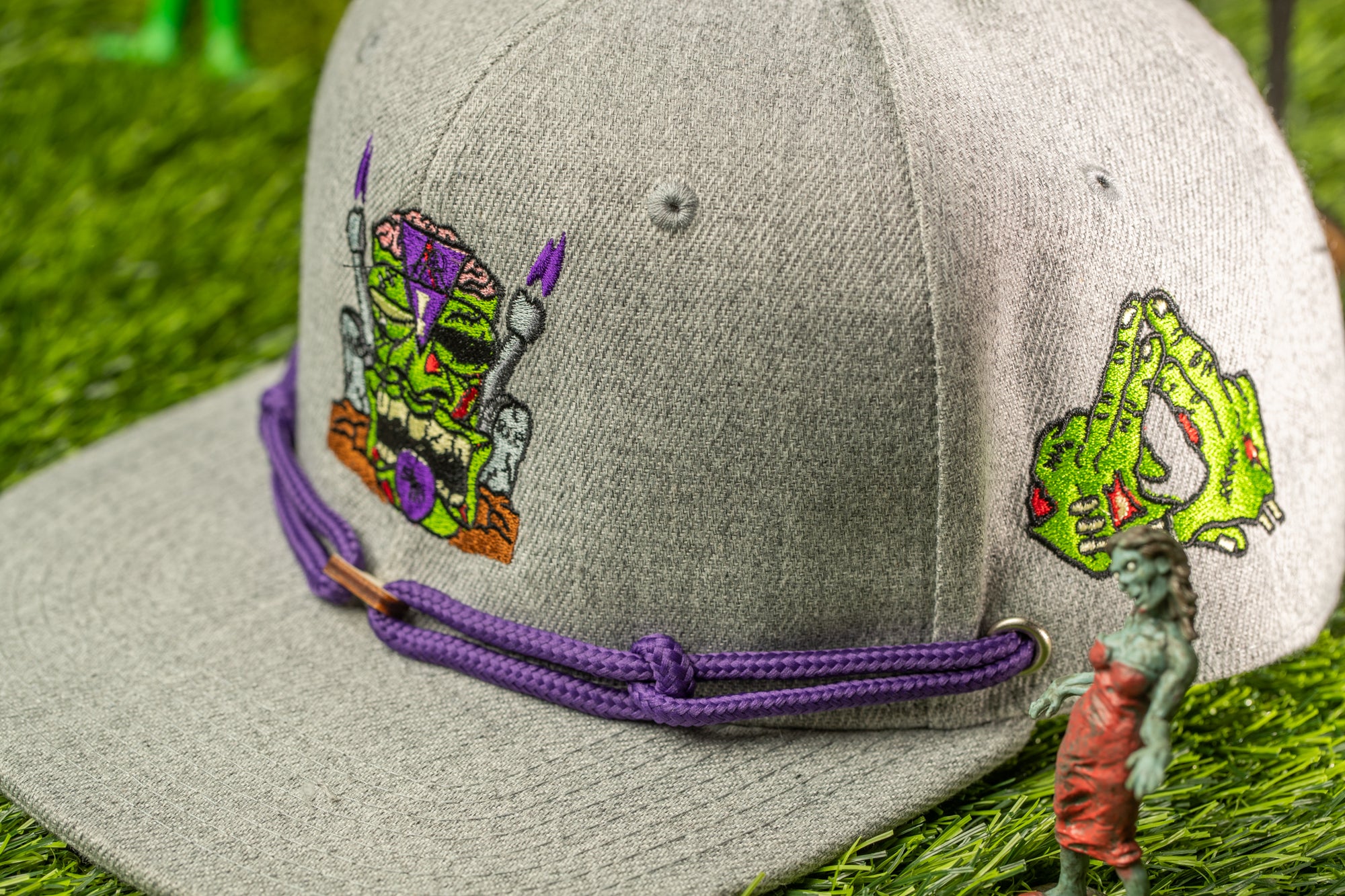 A gray baseball cap with purple and green embroidered designs, including a skull and crossbones, and a small green toy figure of a zombie.