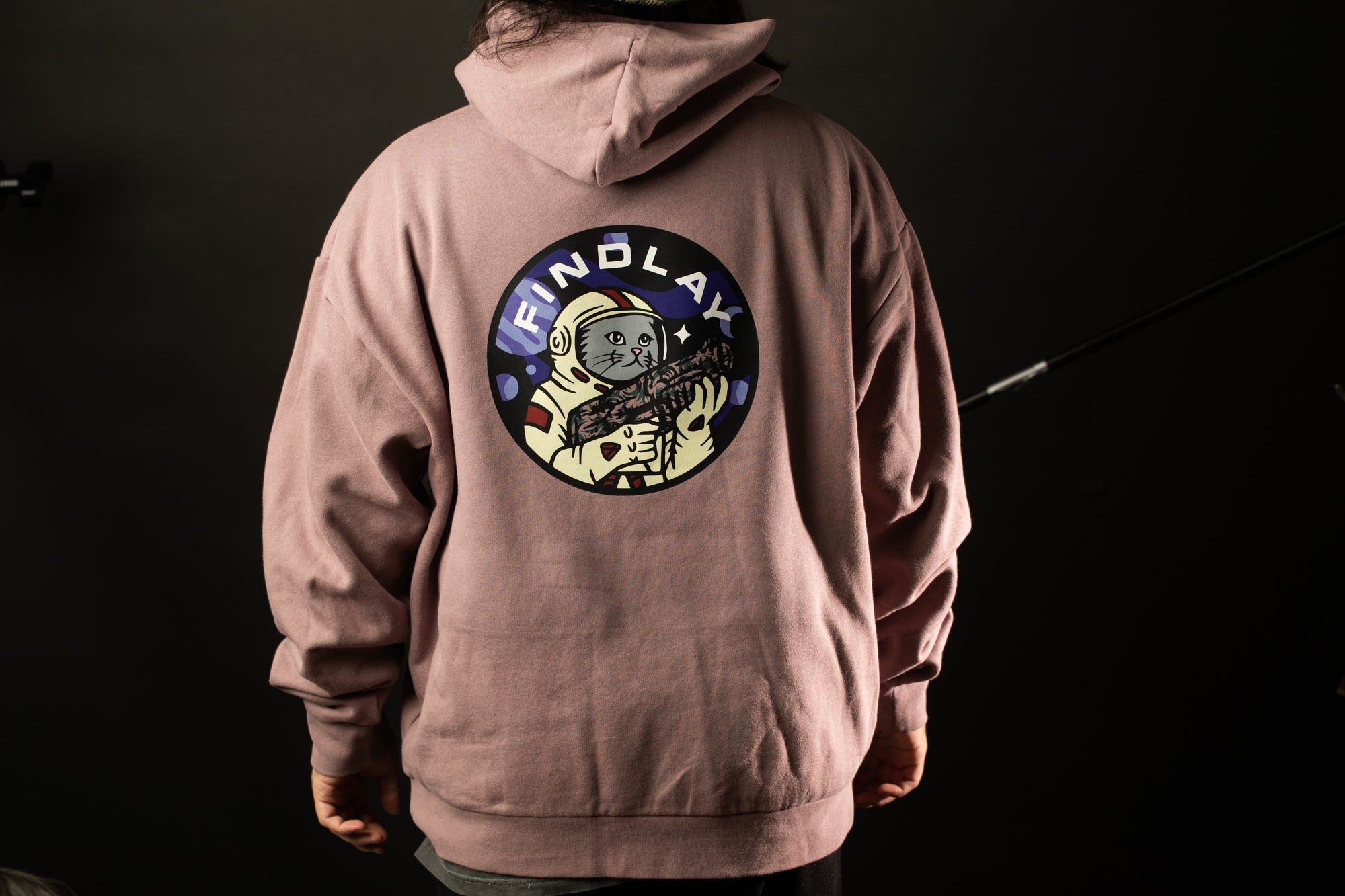 A person wearing a pink hoodie with a graphic of a person in a spacesuit and the text "FIND LAY" printed on the back.