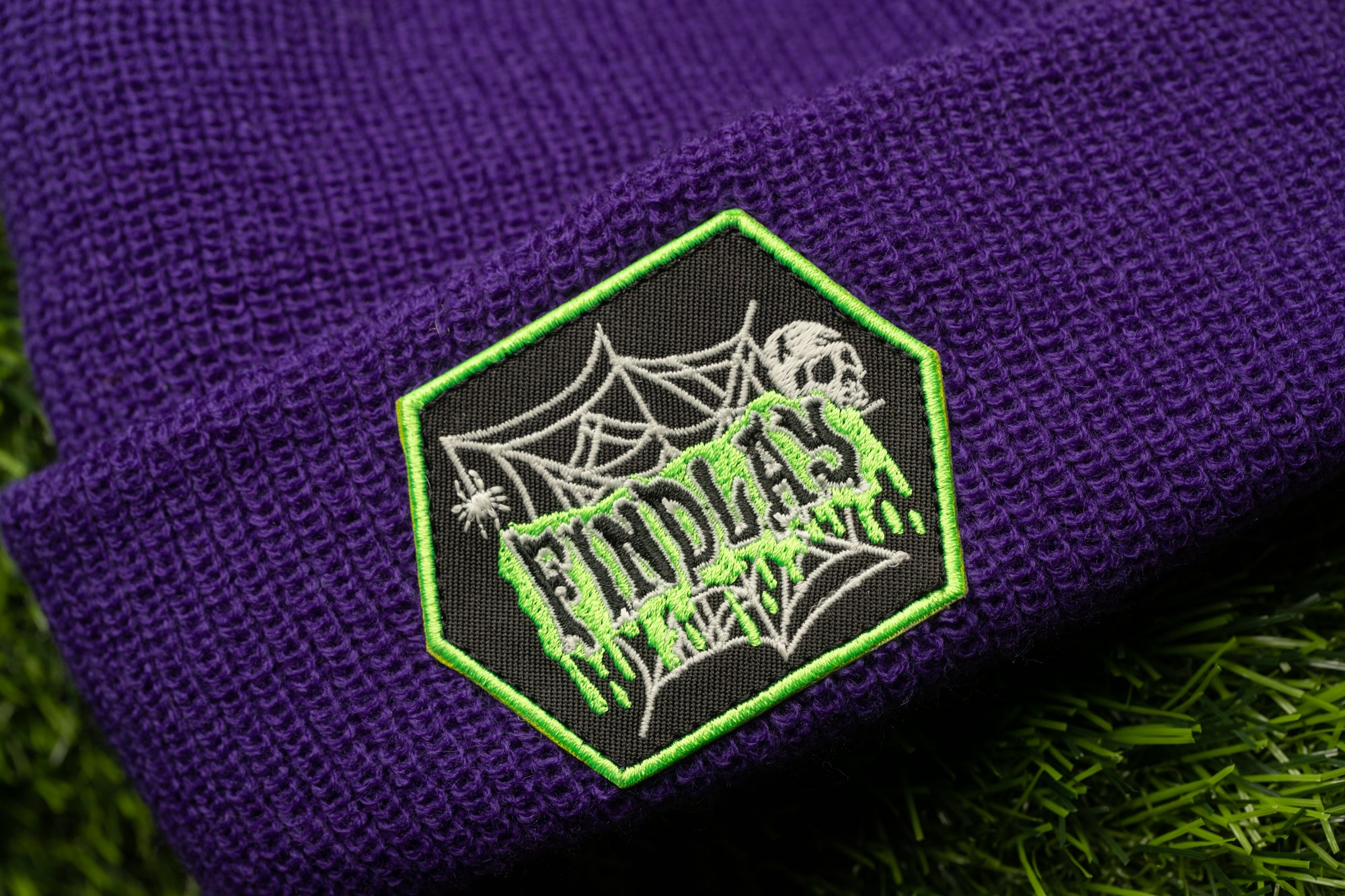 A purple knit beanie with a green hexagonal patch on it, featuring a spider web design and the word "FINDAS" written in white text.