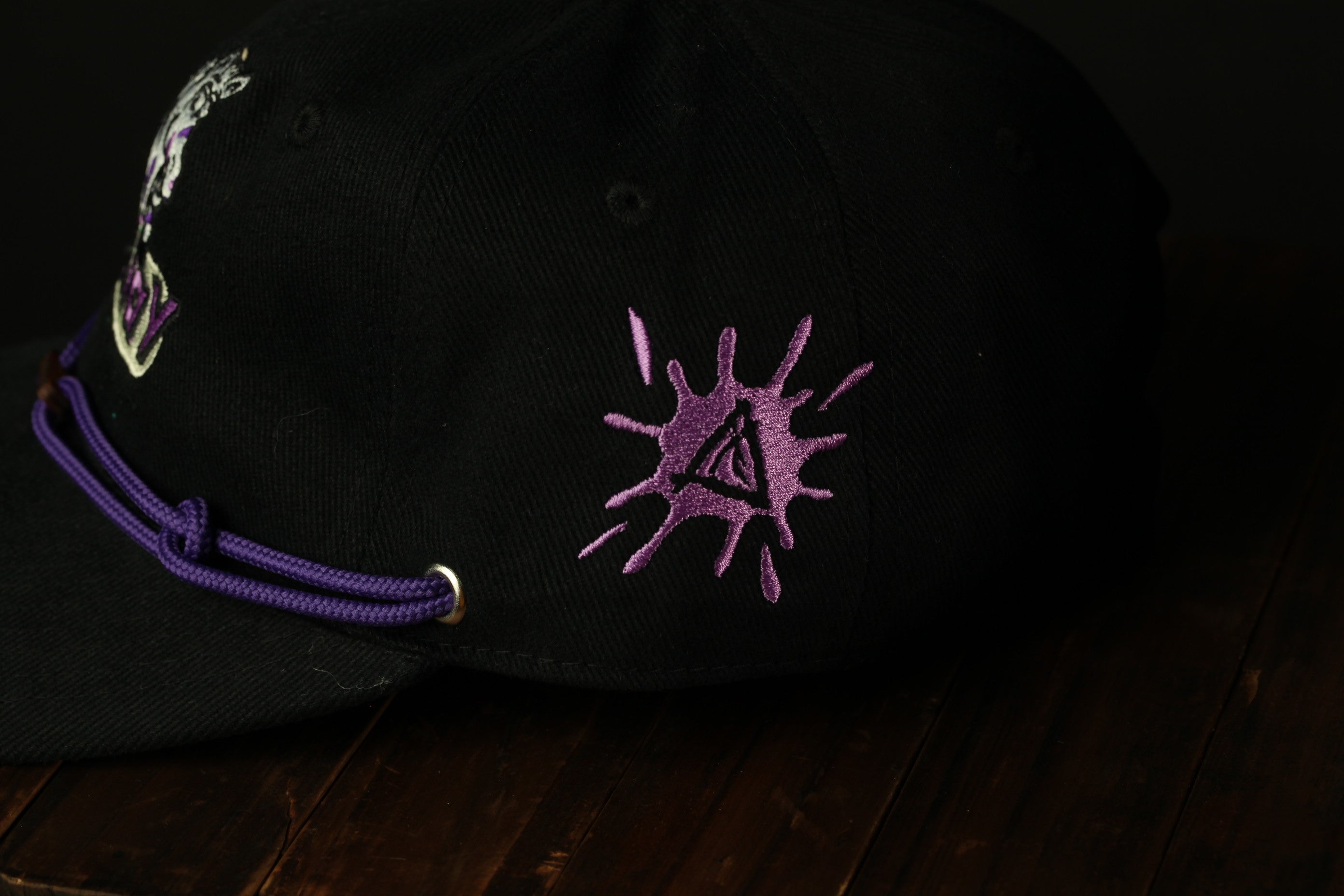 A black baseball cap with purple embroidered designs, including a splash-like pattern and a symbol, is shown on a wooden surface.