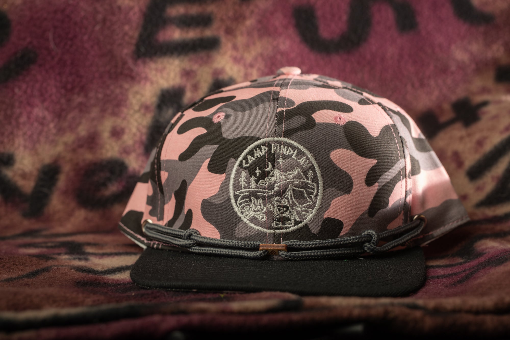A camouflage-patterned baseball cap with a circular logo on the front, resting on a patterned fabric background.