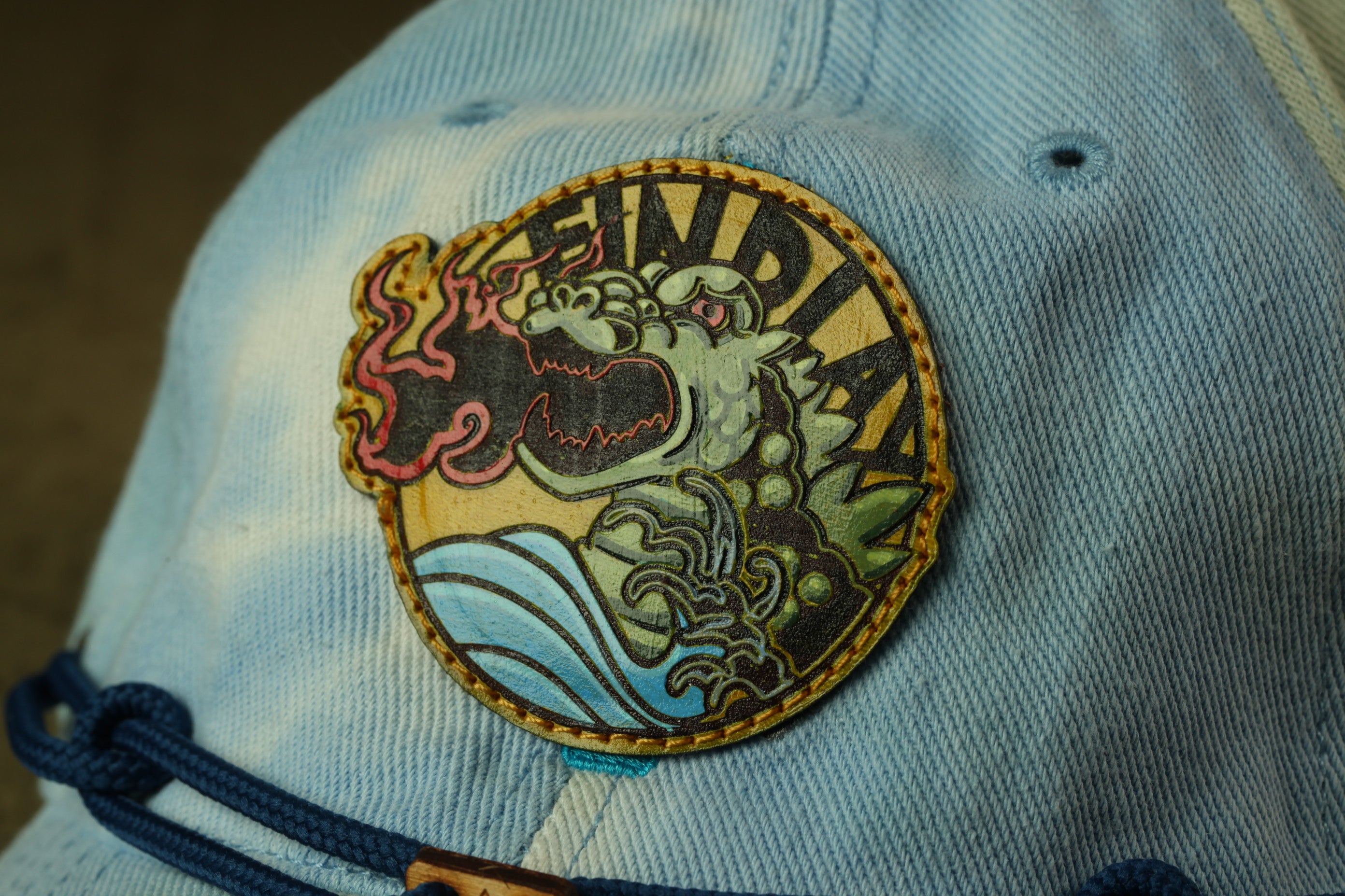 A close-up of a blue denim cap with a circular patch featuring a dragon design and the word "FIND".